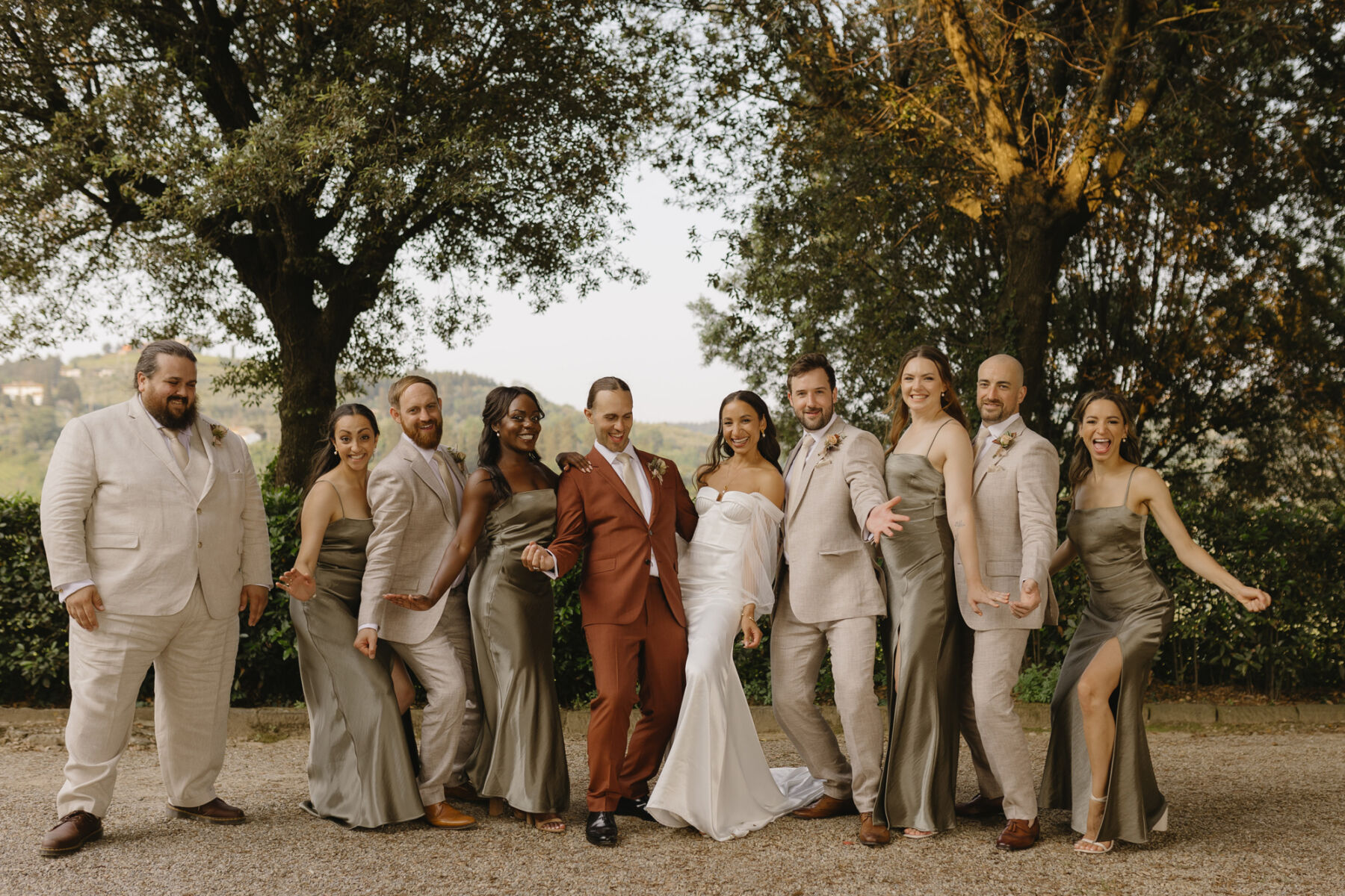 A playful line up at a modern Italian wedding. In the centre, the bride and groom - she wears an Alena Leena wedding dress with detachable tulle sleeves, groom wears a dark orange Paul Smith suit. The bridesmaids wear light green satin dresses by Bec & Bridge and teh groomsmen wear pale suits.