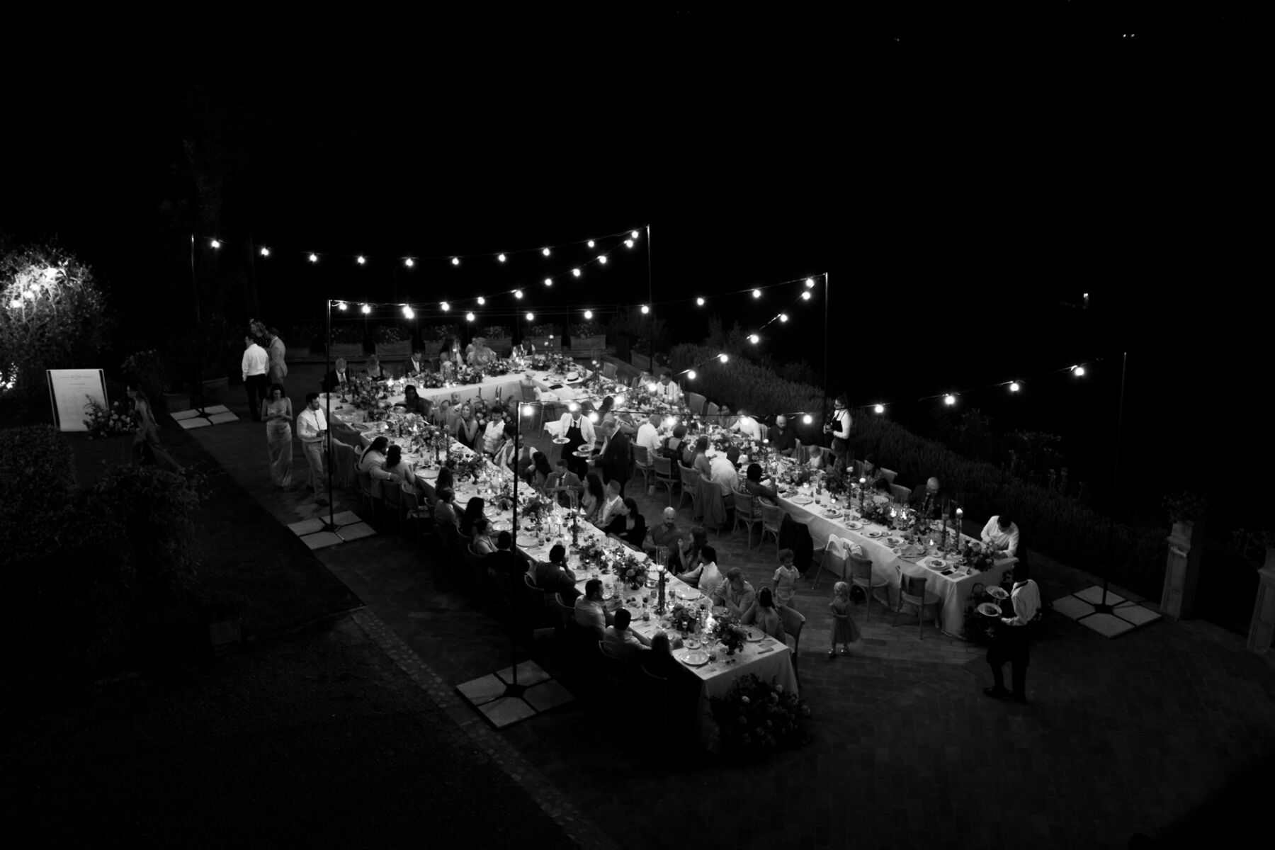 Black and white photograph of festoon lights hanging over a long table set up for an outdoor wedding reception in Italy.