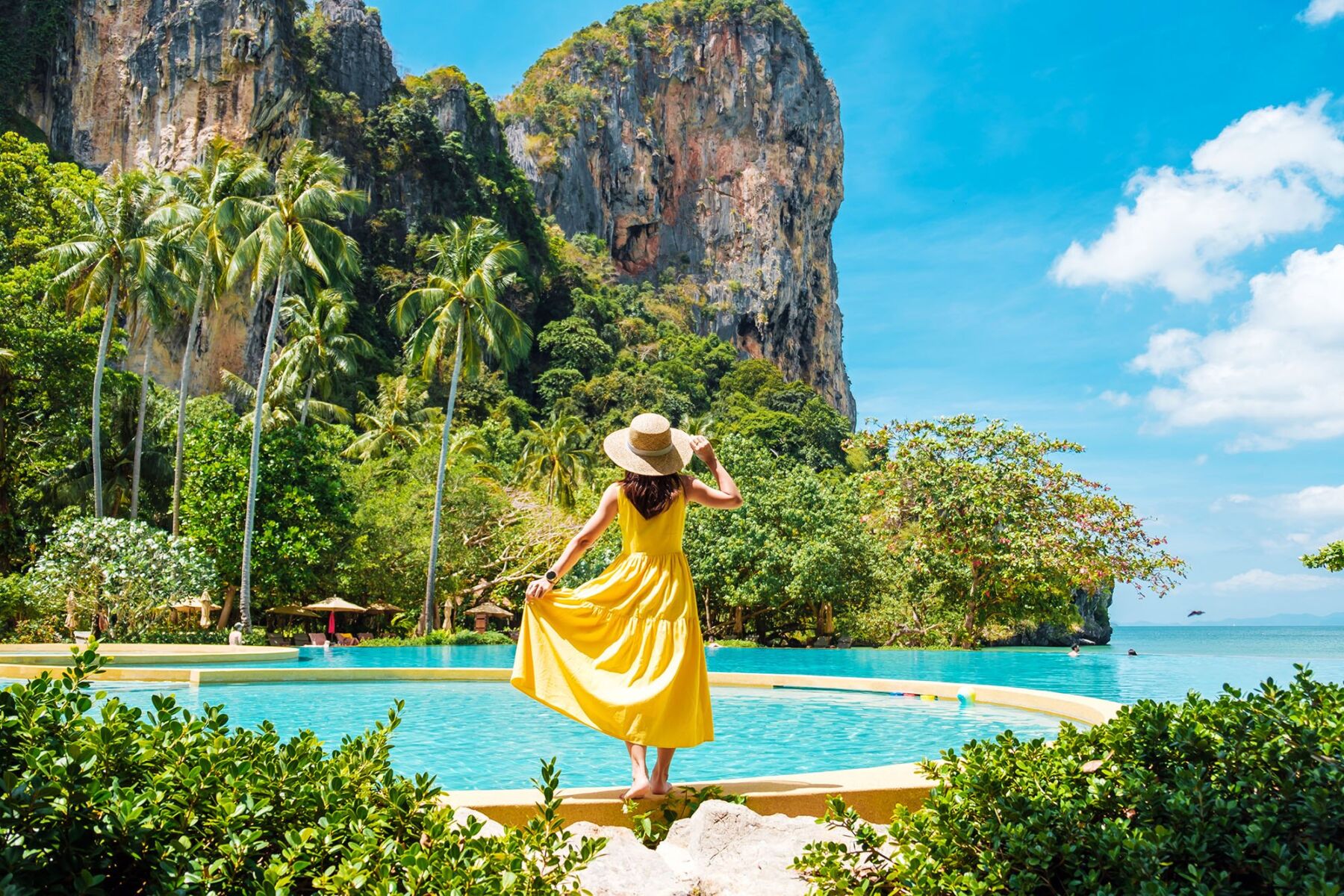 Buy Our Honeymoon - the modern way to plan your honeymoon. Woman in a yellow dress standing by a swimming pool by the sea, surrounded by tall rocks and palm trees.