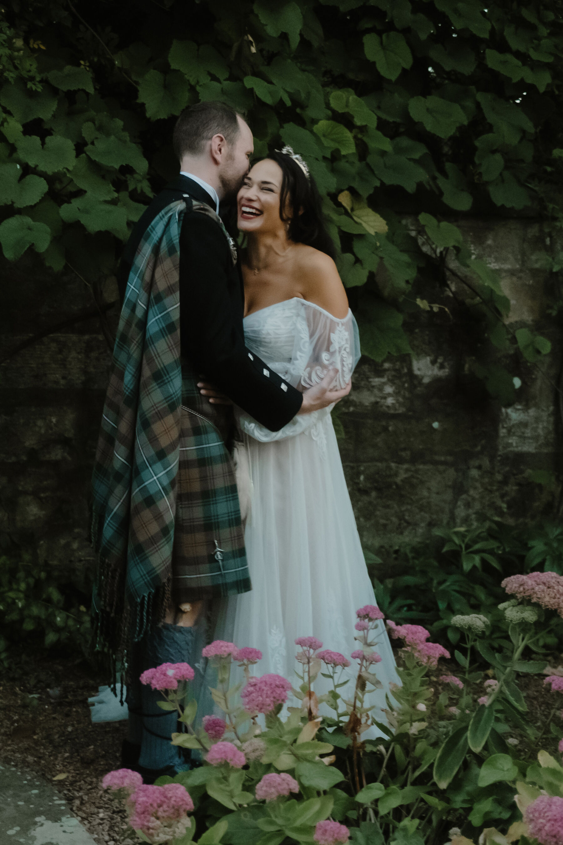 Groom wearing a green and blue tartan fly plaid and embracing his joyful smiling bride who wears an off the shoulder, boho style wedding dress by Willowby by Watters. The couple are standing next to a wall, trees and pink flowers in the grounds of the Cambo Estate, St Andrews, Scotland.