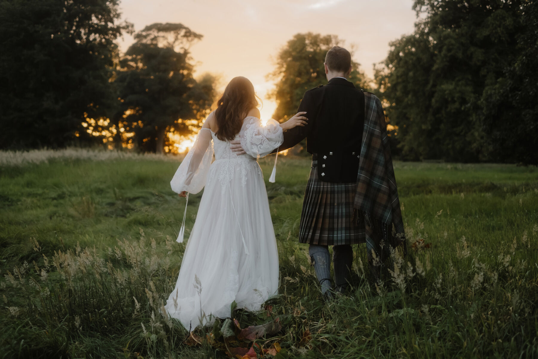 Golden hour shot of bride and groom walking outside. Photograph taken from behind - groom wears a kilt in greens and blues. Bride wears a bohemian inspied wedding dress by Willowby Watters.