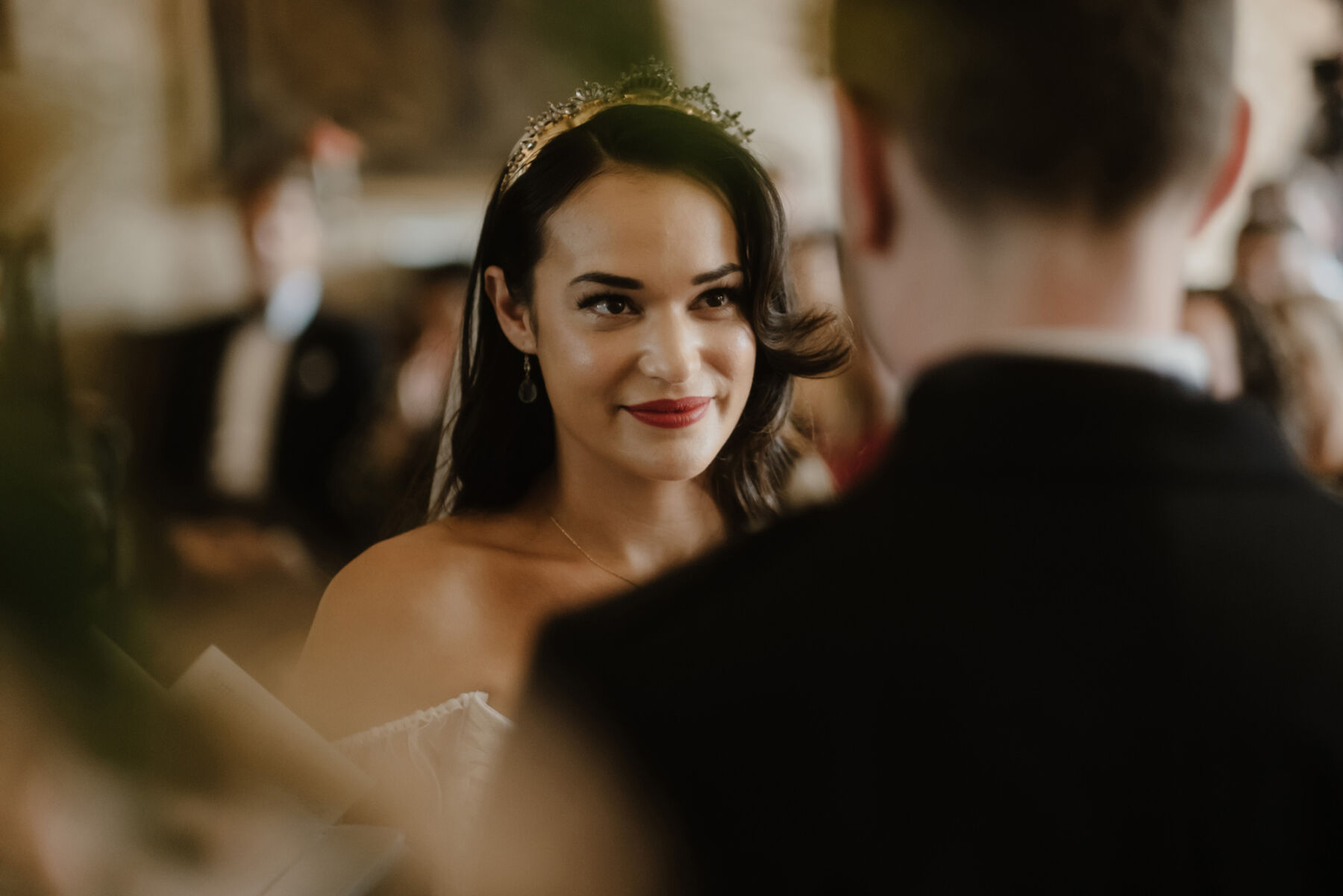 Bride looking lovingly towards her groom. She has long dark her, wears red lipstick, a gold Lelet headpiece and a bohemian off the shoulder gown by Willowby by Watters.