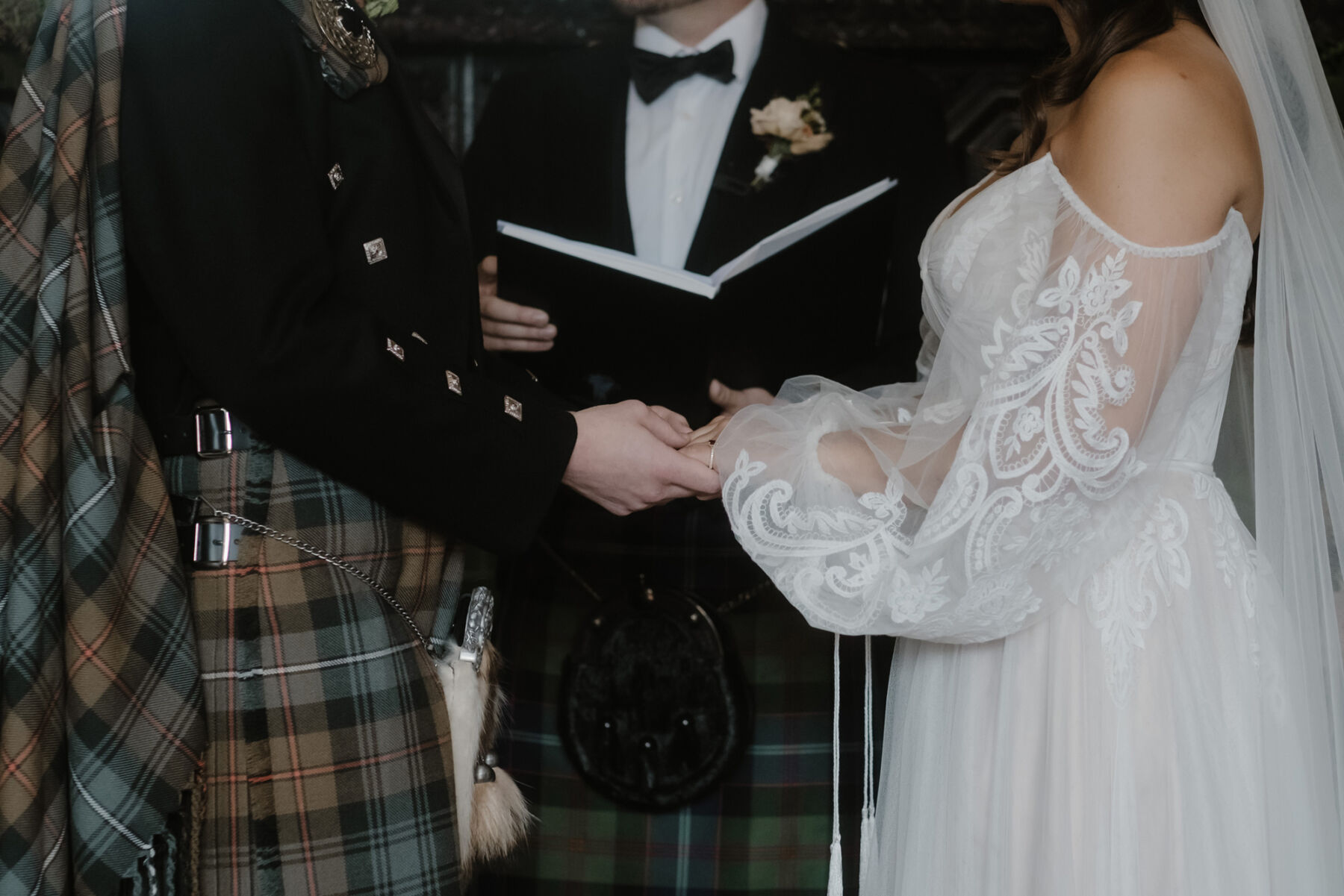 Image focussing on the bride and groom holding hands during their wedding ceremony at Cambo Estate. Groom in a kilt, bride wears an off the shoulder gown by Willowby by Watters. 