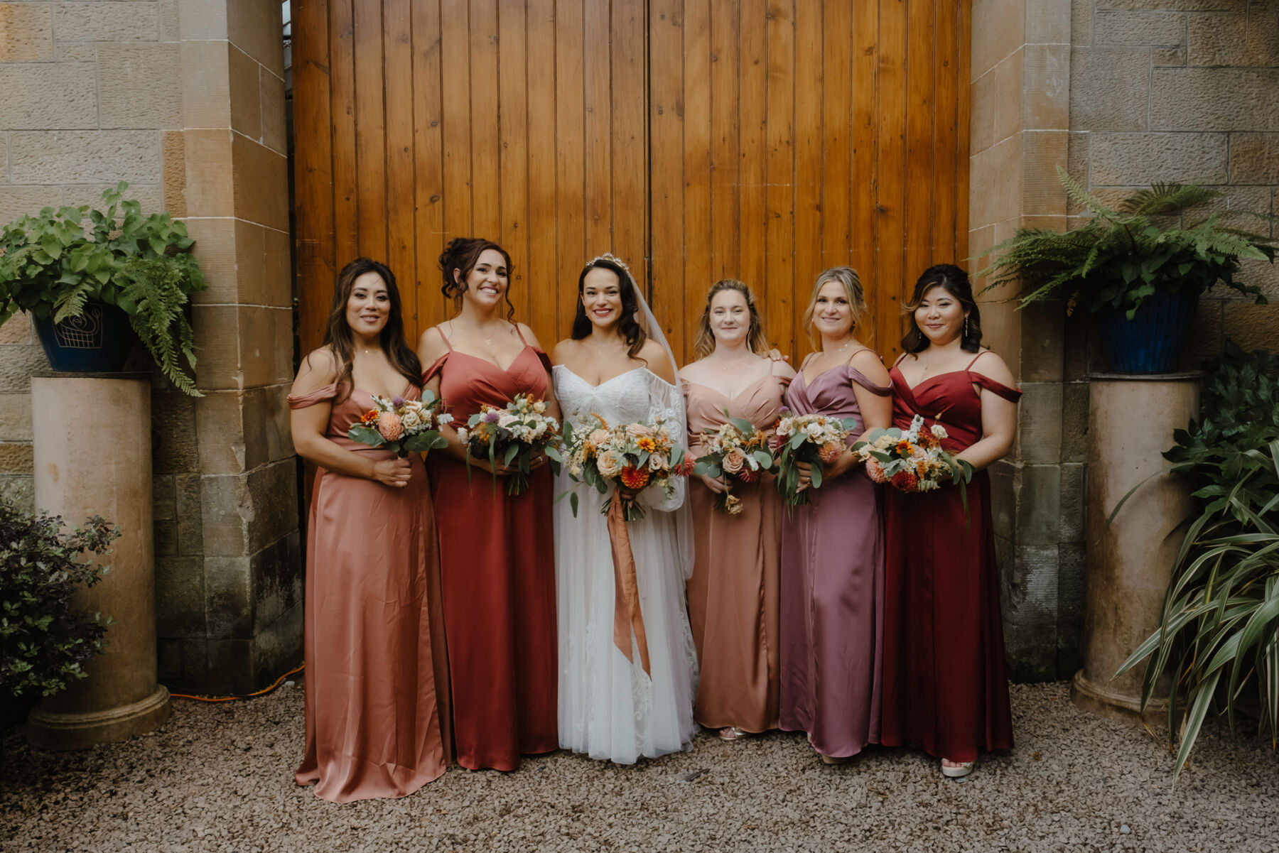Bridesmaids wearing orange, coral, peach and pale pink wedding dresses by Azazie. The bridesmaids carry Autumn bouquets in soft peach, coral and pink.