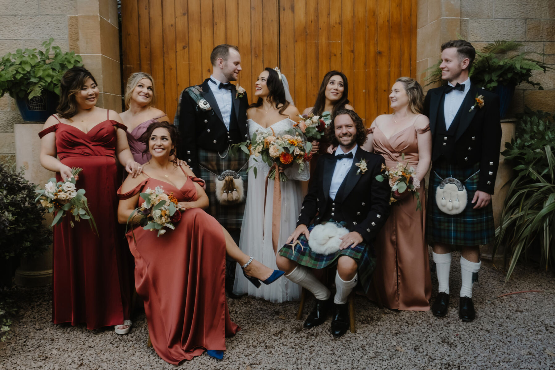 Bridal party at Cambo Estate Scotland. Groom & groomsmen in kilts with blue and green tartan. Bridesmaids in Autumn shades of burnt orange, coral, red and peach. Bride wears a boho style gown by Willowby by Watters.
