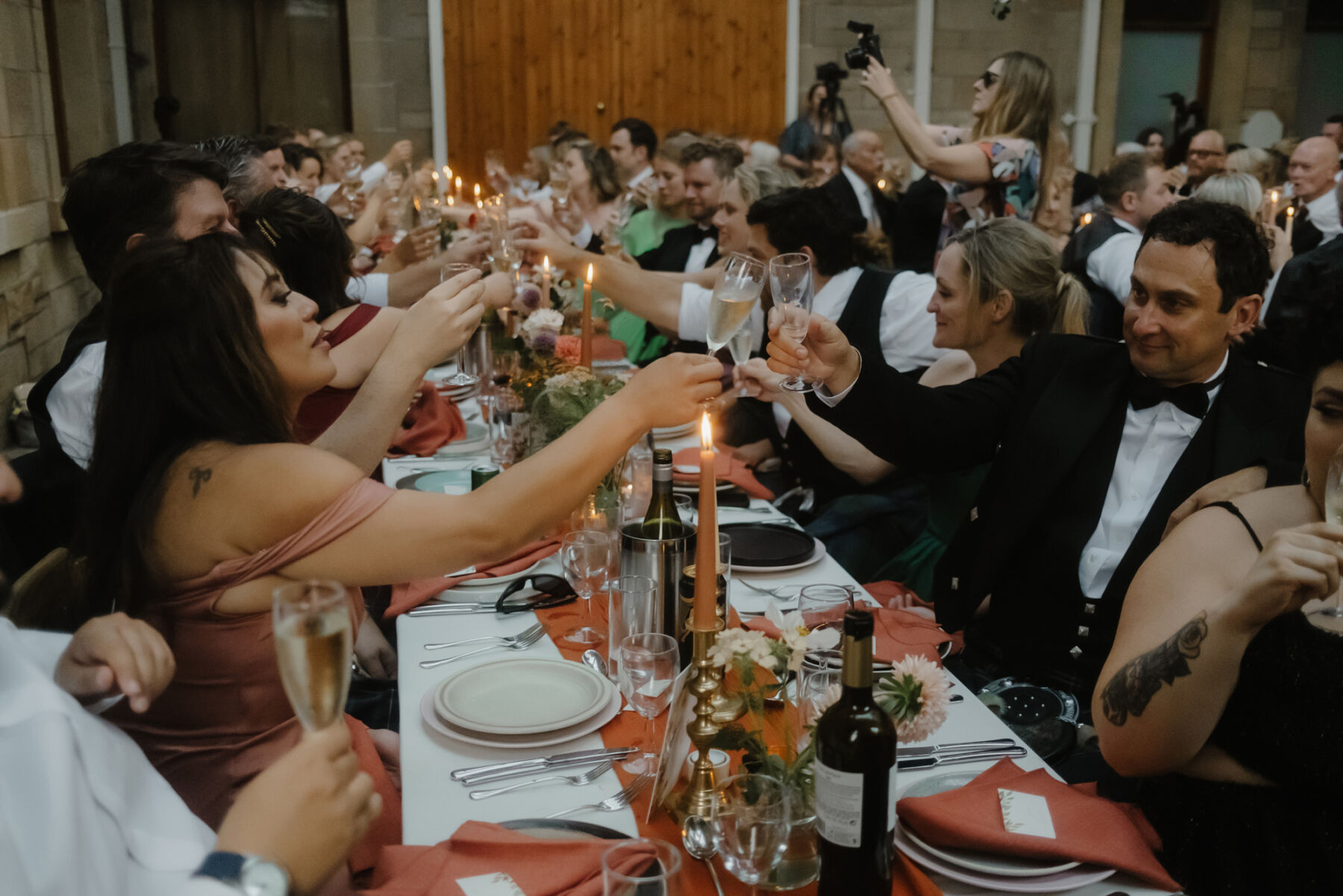 Guests chinking their champagne glasses across a candle lit table at Cambo Estate in Scotland. Burnt orange napkins and pale pink dahlia.