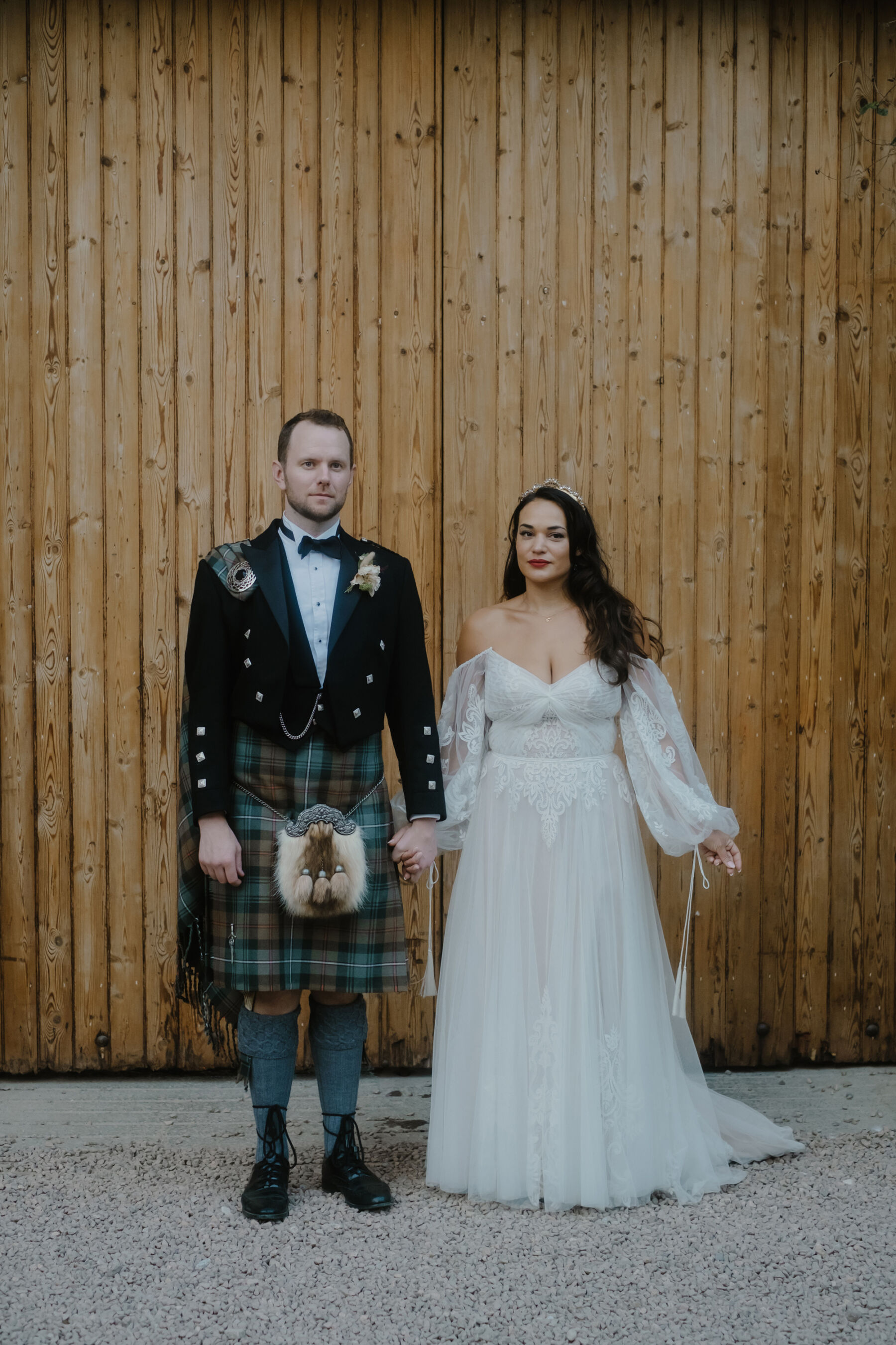 Groom wearing a green holding hands. Groom wears a green and blue tartan kilt and fly plaid with a sporran. Bride wears an off the shoulder wedding dress by Watters. She has long dark loose hair, red lipstick and pale pink nails.