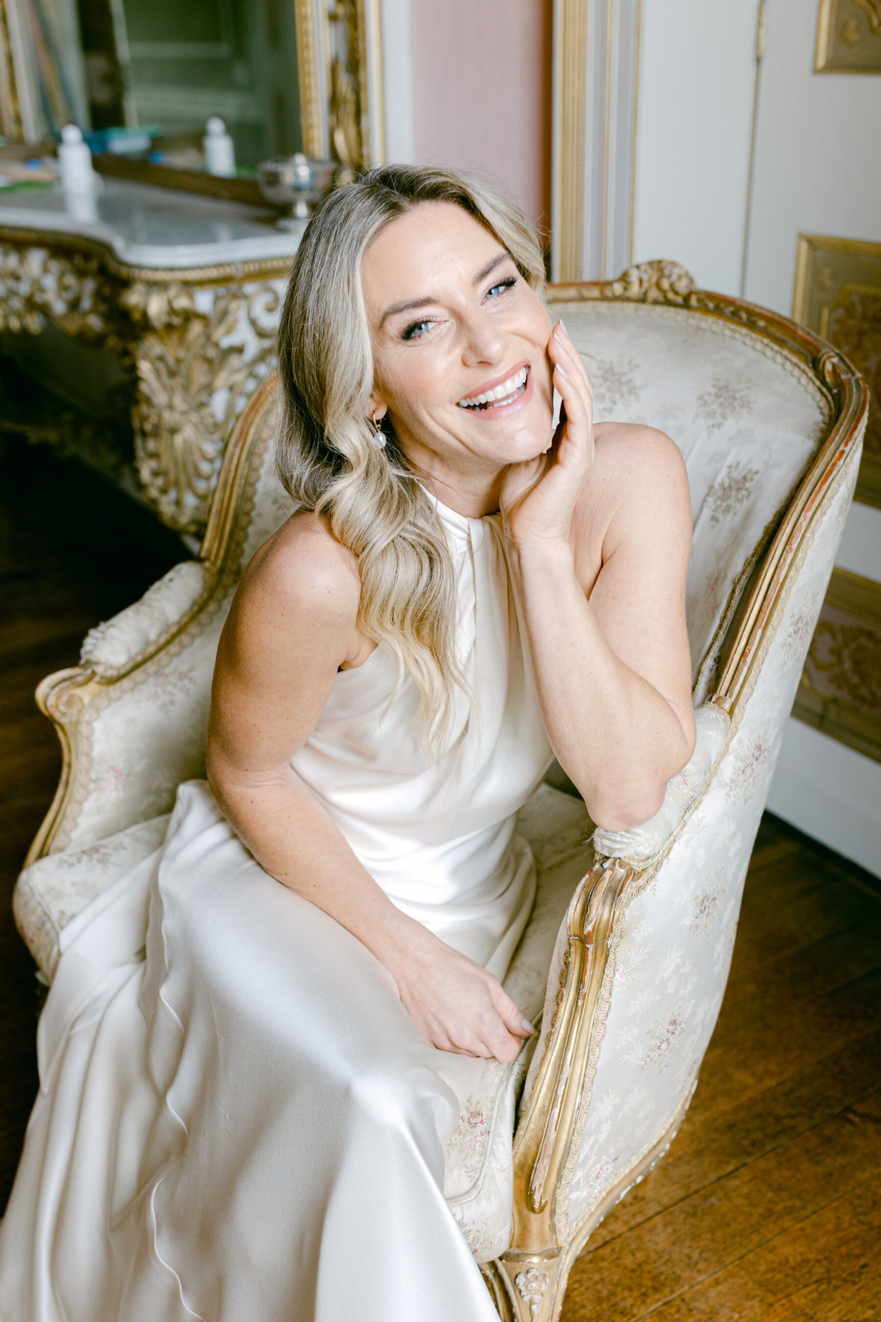 Laughing happy bride seated, wearing a silk halterneck wedding dress by Halfpenny London.