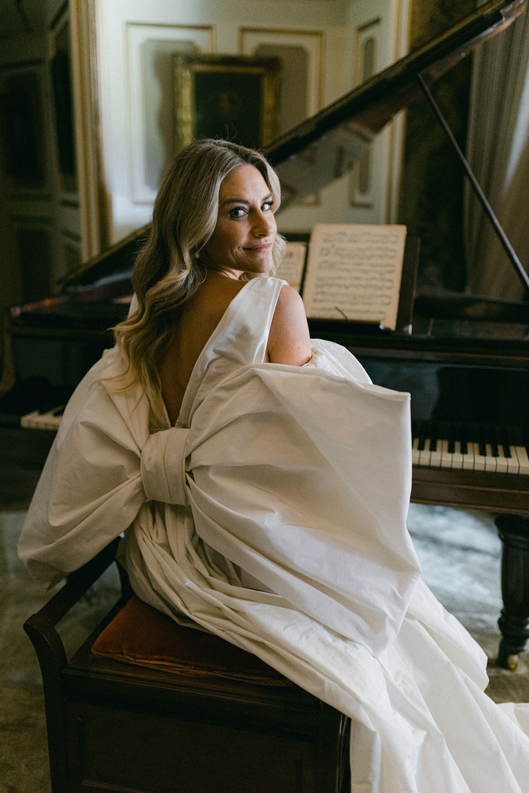 Big bow.Wedding dress with a huge statement bow at the back, by Halfpenny London. Bride is sat at the piano and turning around to to look at the viewer.