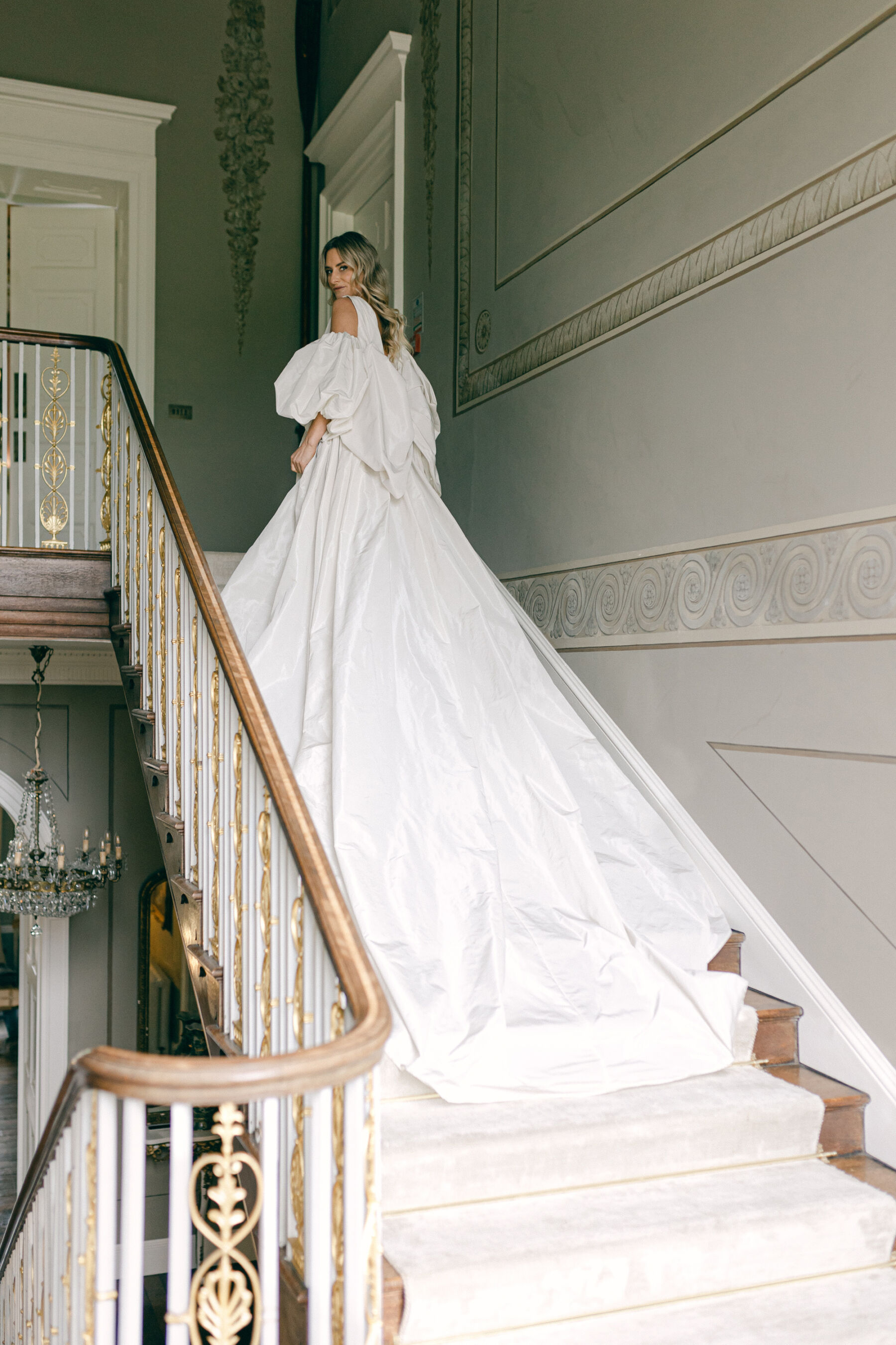 Bride wearing a dress with a long train is standing on the stairs at Avington Park in Hampshire 
