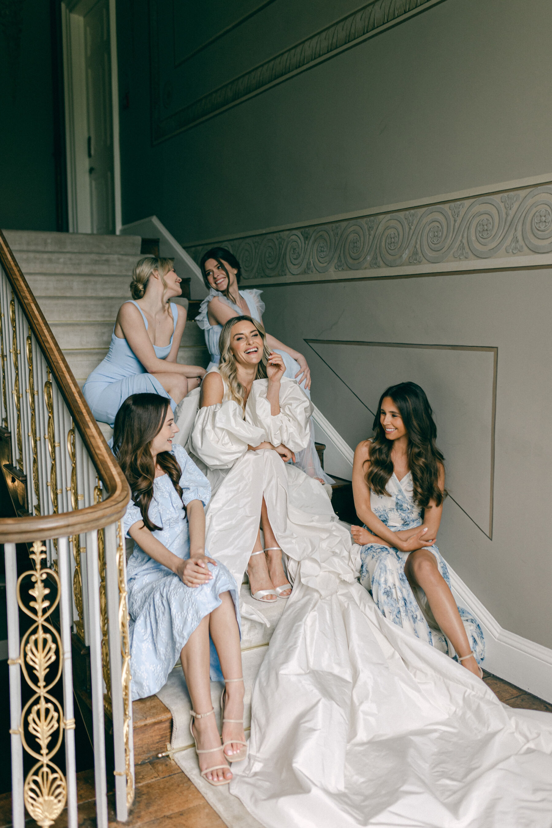 Bride and bridesmaids sat on the sweeping staircase at Avington Park, laughing and jolly. The bride wears a Halfpenny London bridal gown full of fashion drama and the bridesmaids wear pale blue dresses.