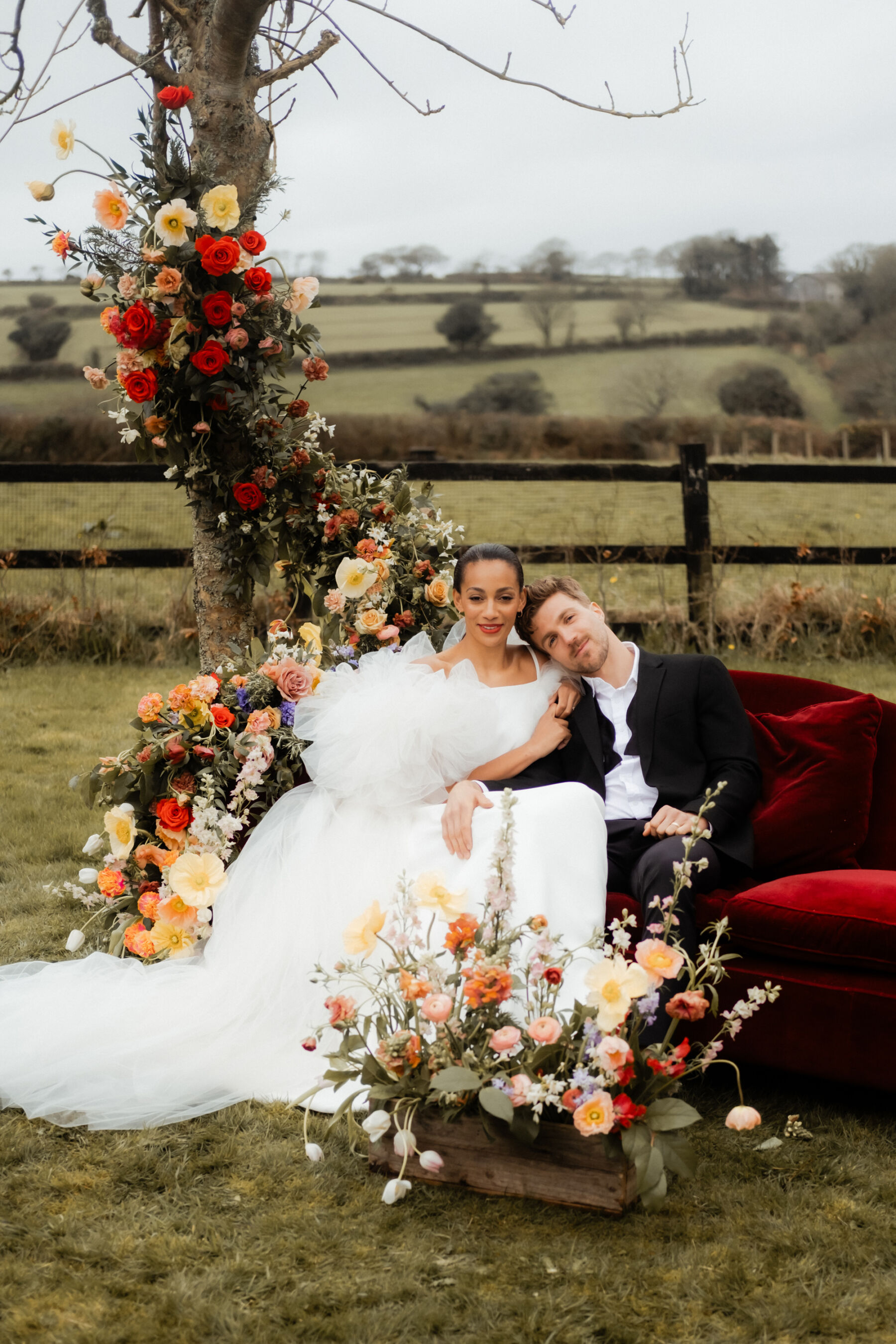 Bride and groom sat on a red plush sofa outside with flowers everywhere. The Cornish Place, Cornwall