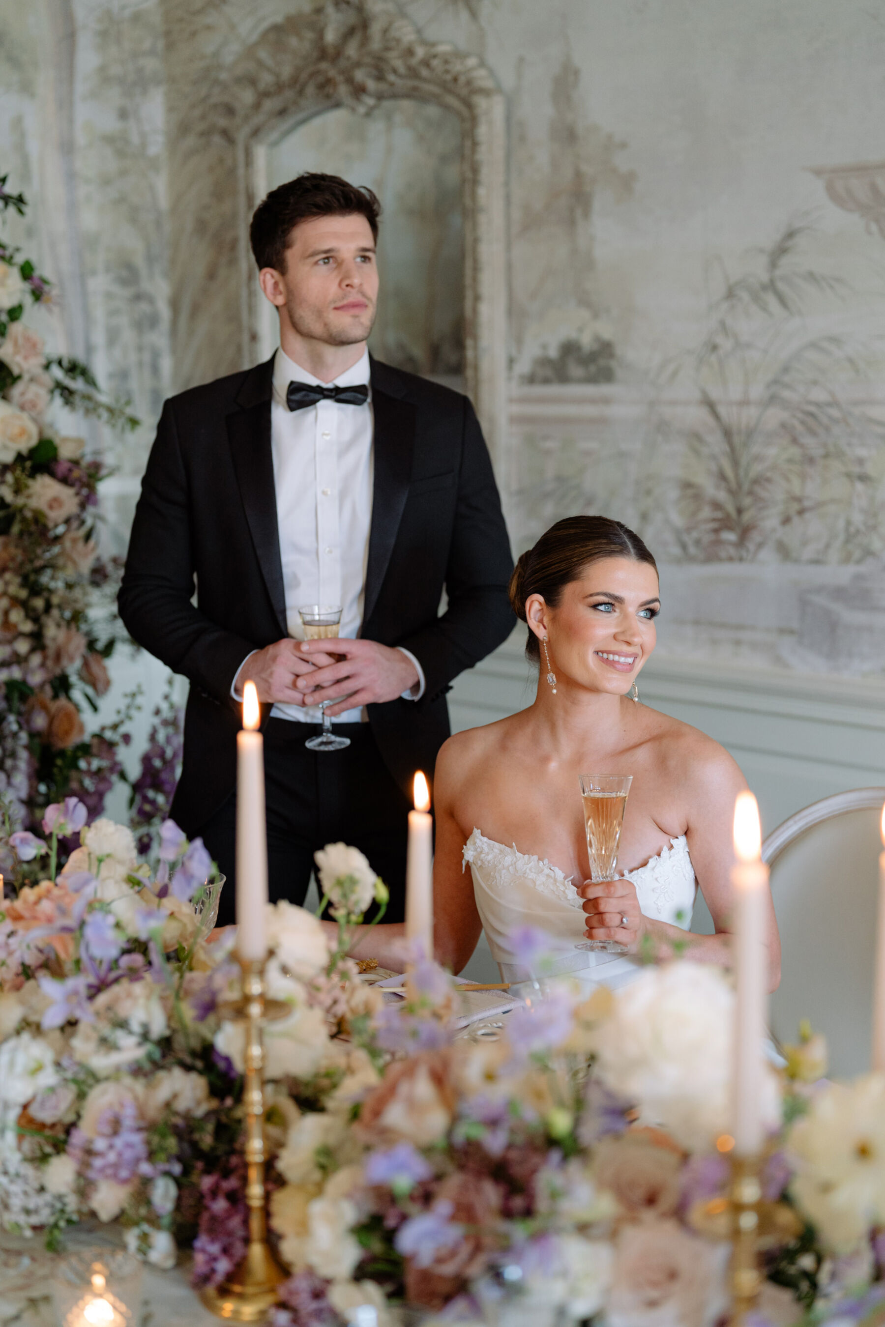 Model bride wearing a modern, strapless Josephine Scott London bridal gown. She is seated next to her standing groom in black tie, next to a table with a display of Bridgerton inspired flowers. Kamila Nowak Photography.