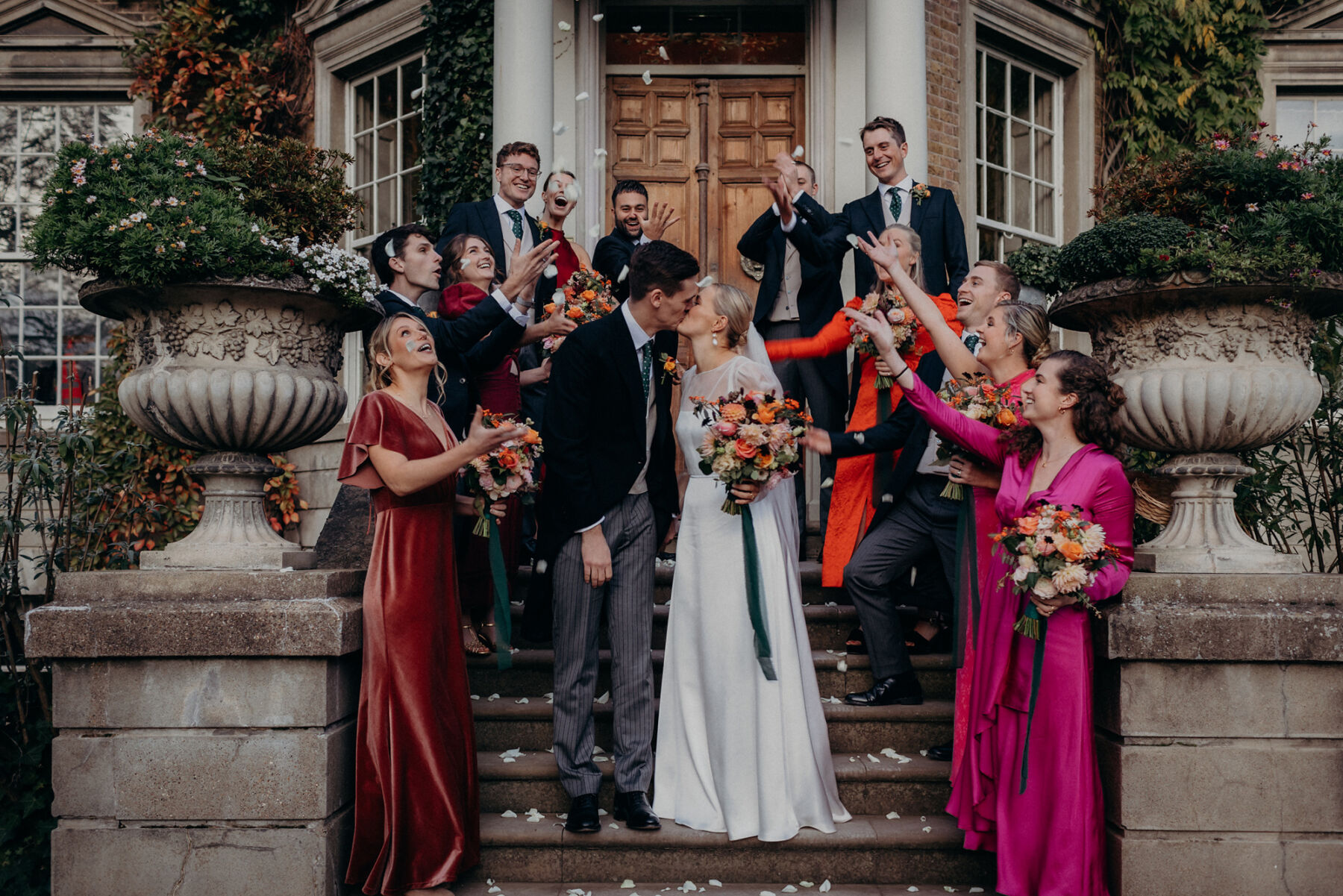Bride and groom standing on the steps at Hampton Court Palace surrounded by the bridal party showering them in confetti. The bridesmaids wear bright pink and red dresses. Victoria Somerset-How Photography.