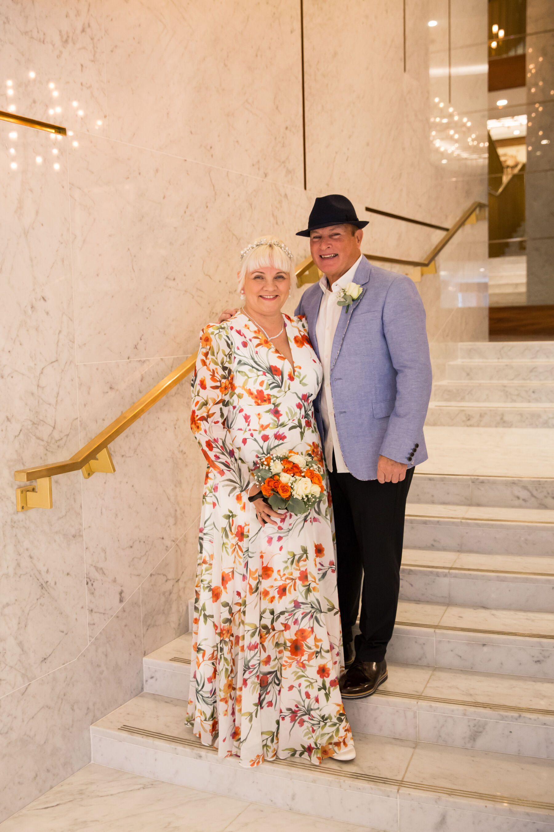 Mature couple on their wedding day. The bride wears a floral dress and carries a small bouquet of orange and cream roses and the groom wears a light blue jacket and fedora hat. 