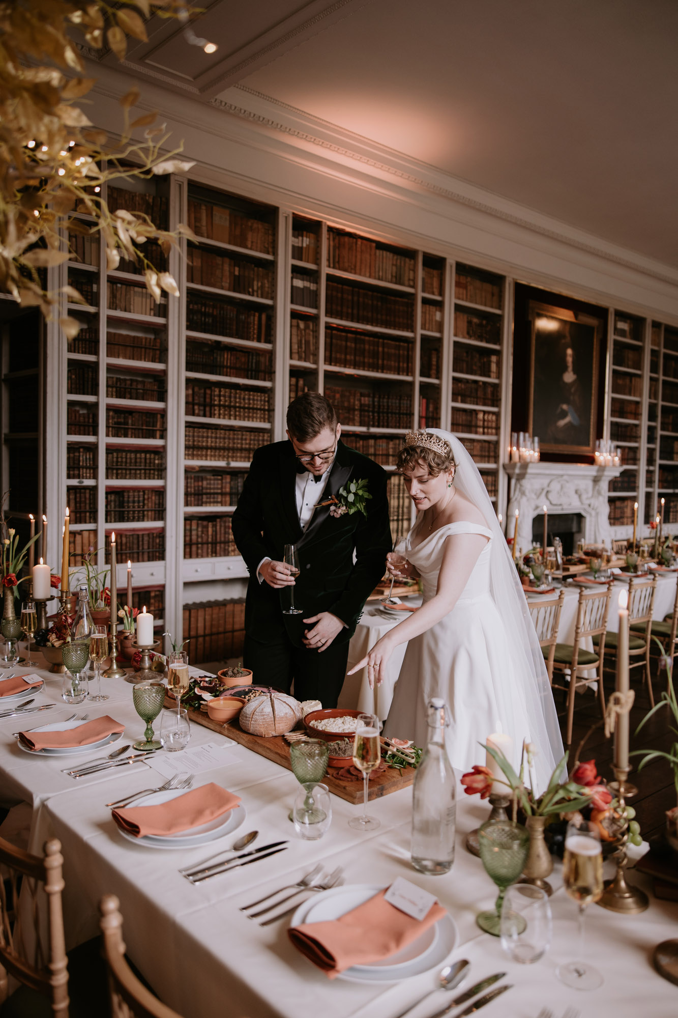 Bride and groom walking around the reception room at St Giles House in Dorset, tables decorated with green wine glasses, pale orange napkins and tall taper candles. Marta Ilardo Photogarphy.