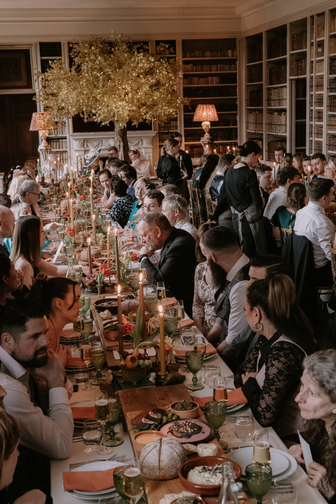 Guests dining at St Giles House wedding venue, Dorset. Long feasting tables with tall taper candles, burnt orange napkins, fresh bread, vegan food. Marta Ilardo Photography.