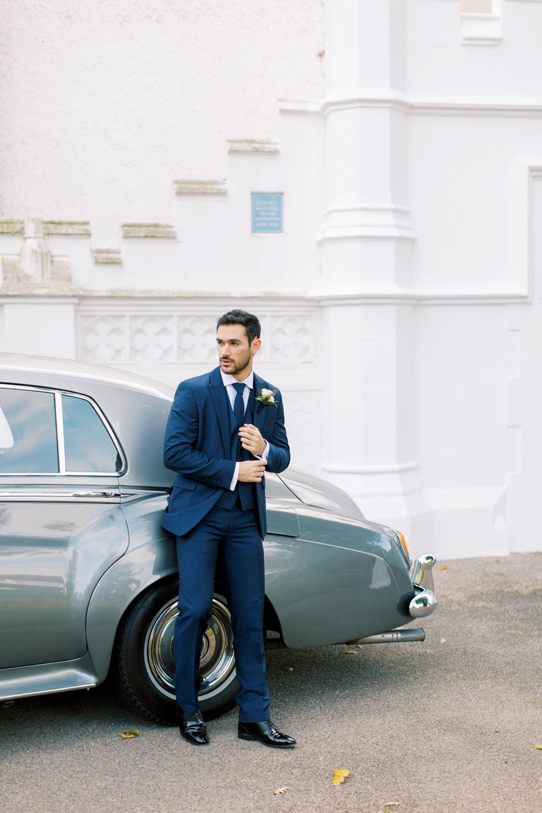 Gothic Architecture Inspired Wedding Styling, at Strawberry Hill House 23 Stylish groom in a blue suit leans against a Rolls Royce that is parked outside Strawberry Hill House & Garden. Helen Warner Photography.