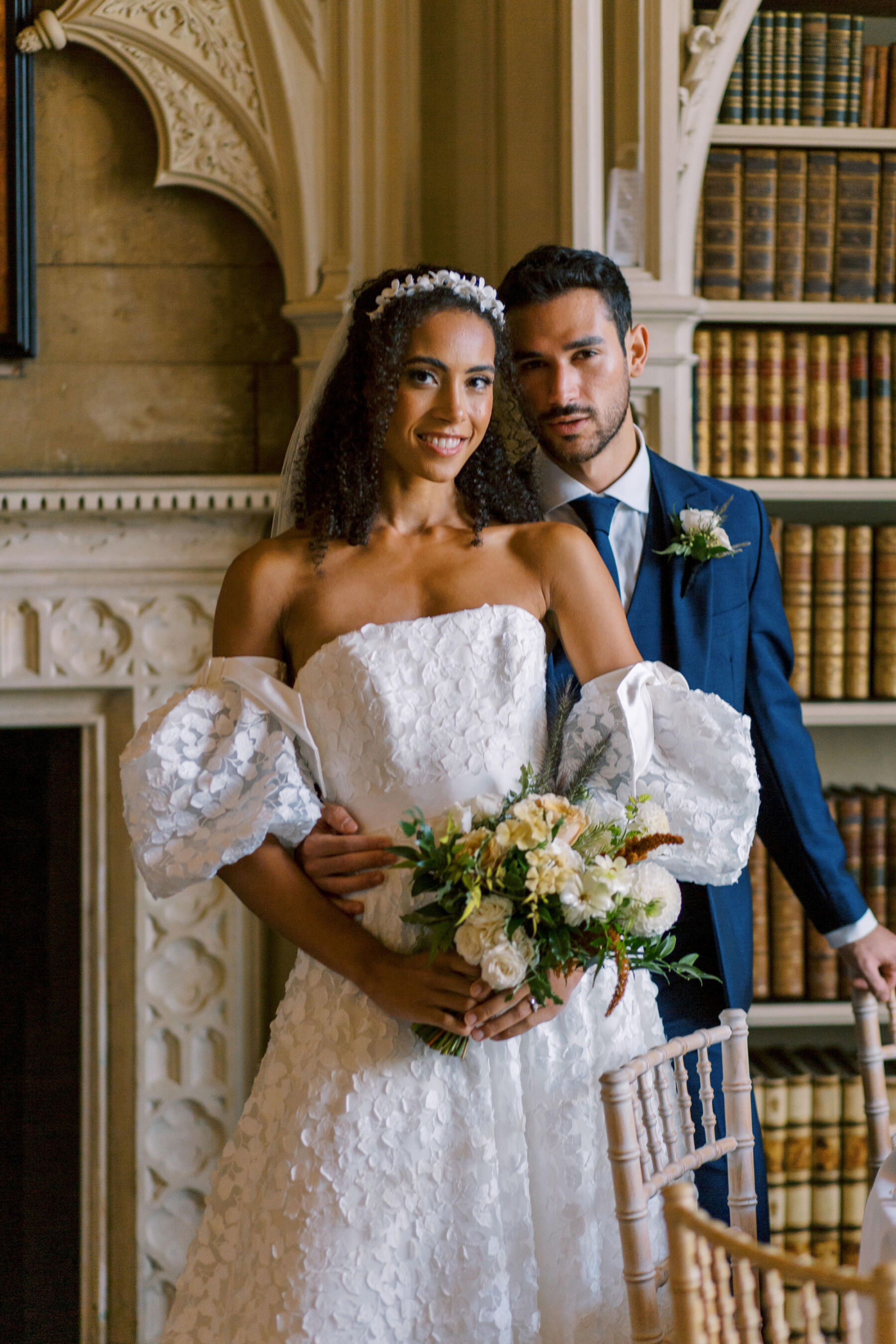 Gothic Architecture Inspired Wedding Styling, at Strawberry Hill House 4 Sassi Holford puff sleeve wedding dress. Bride holds a modern wedding bouquet on neutral and toffee tones. Groom stands behind her clutching her waist wearing a modern blue suit. The backdrop is the inside of Strawberry Hill House & Garden. Helen Warner Photography.