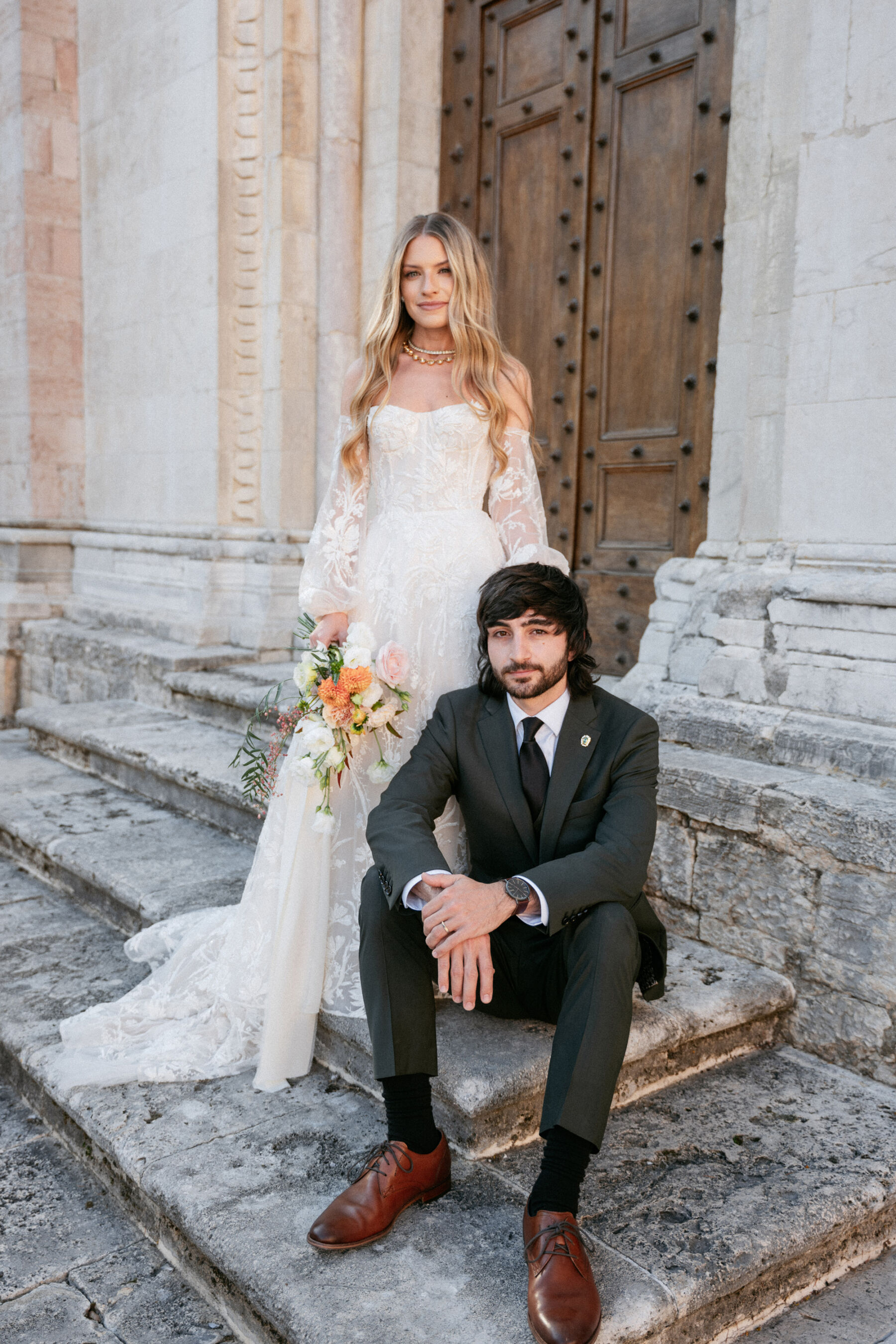 Mikaela in Alena Leena For Her Elegant Italian Wedding at Villa Fattoria di Vibio 8 Bride with long blonde wavy hair stood on the steps outside an Italian church, her husband seated on the steps. She wears an Alena Leena wedding dress with removable sleeves and carries a bright medium sized bouquet. Photography by Stefano Santucci.