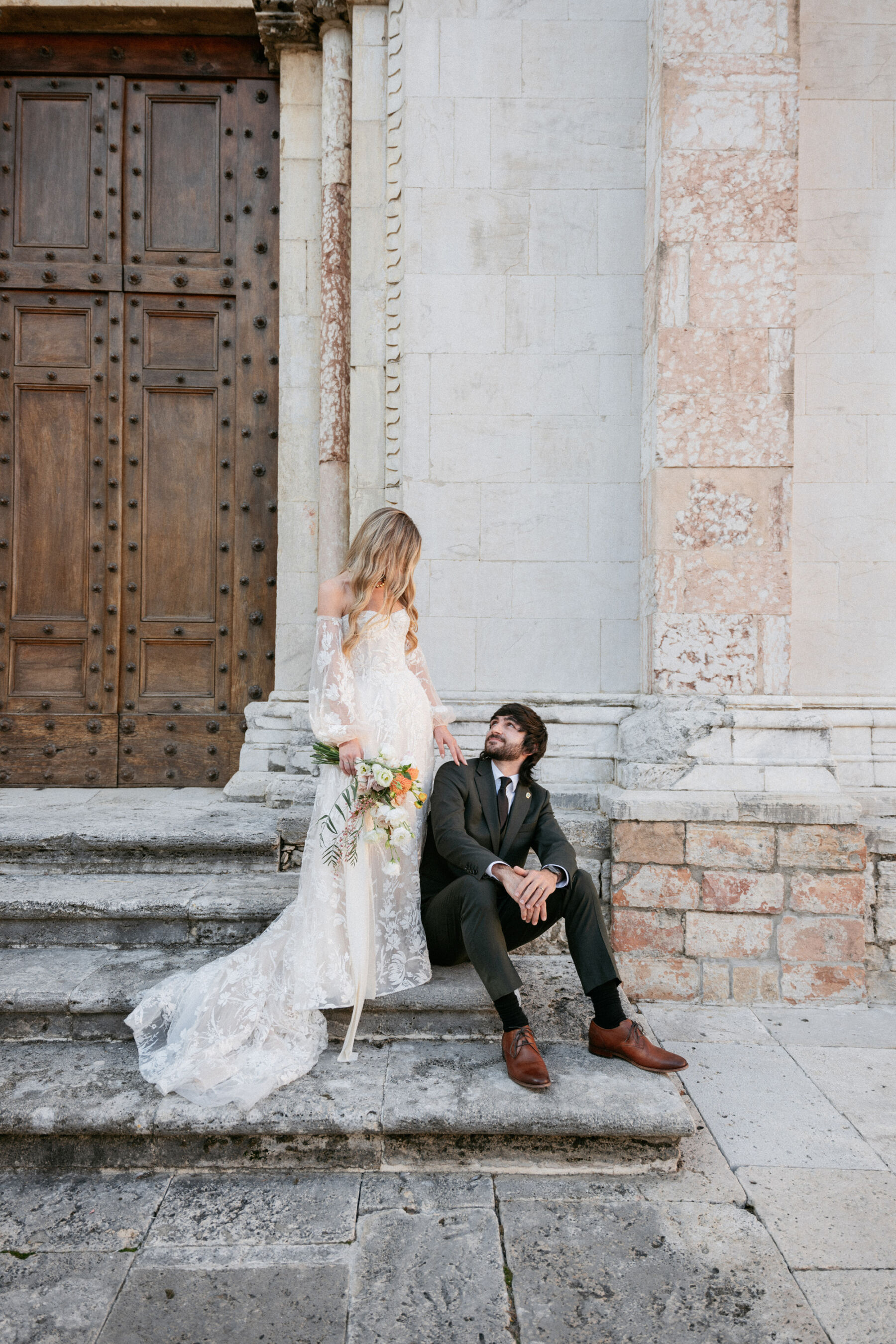 Mikaela in Alena Leena For Her Elegant Italian Wedding at Villa Fattoria di Vibio 36 Bride with long blonde wavy hair stood on the steps outside an Italian church, her husband seated on the steps. She wears an Alena Leena wedding dress with removable sleeves and carries a bright medium sized bouquet. Photography by Stefano Santucci.