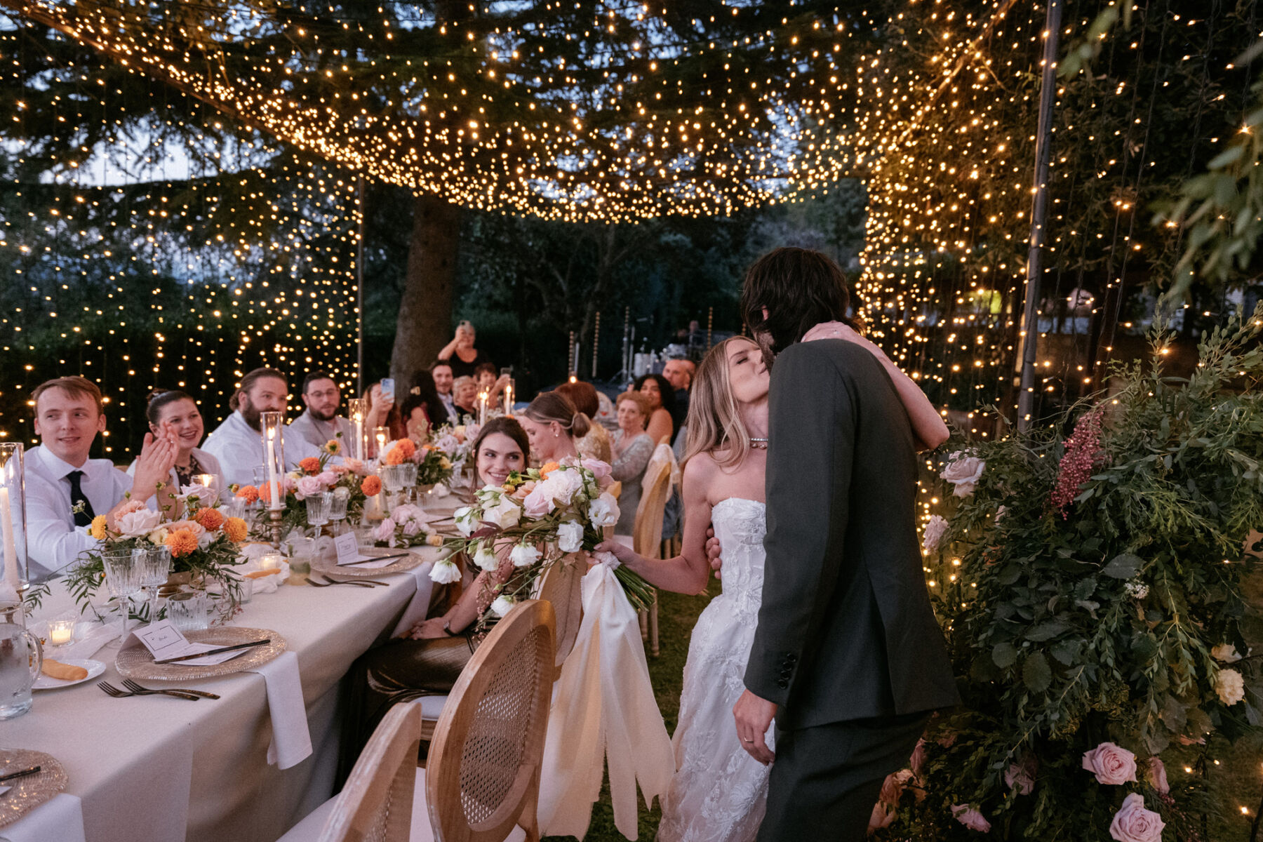 Mikaela in Alena Leena For Her Elegant Italian Wedding at Villa Fattoria di Vibio 44 Bride and groom kissing before being seated at their Italian wedding reception. The reception table is positioned beneath a canopy of twinkly lights and guests are turning to smile at them. The bride holds and elegant bouquet with a large silk ribbon. Photography by Stefano Santucci.