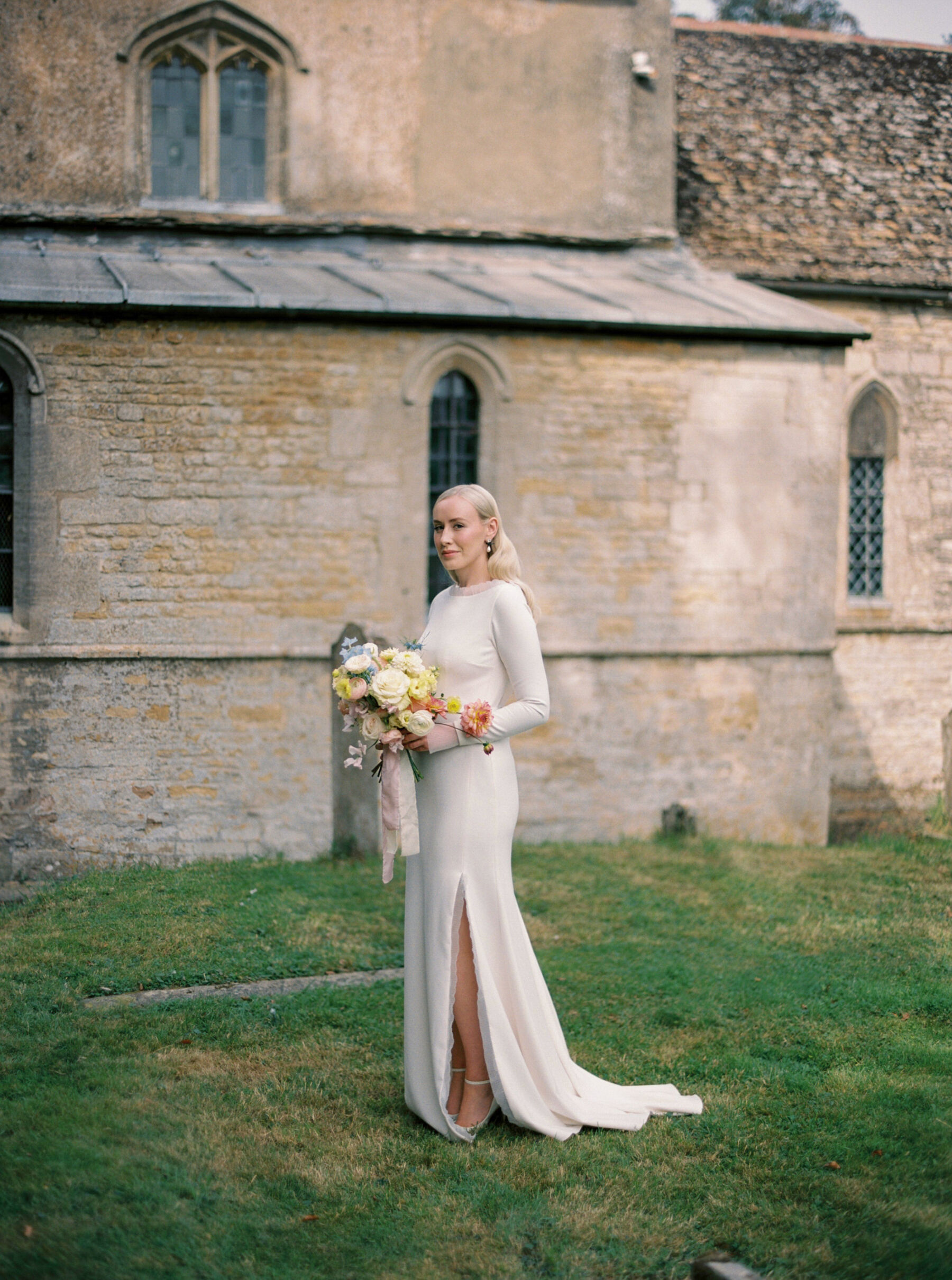 Annie & Harry's Effortlessly Glamorous, Colourful & Modern Double Wedding Weekend 2 Beautiful bride wearing a bespoke Stephanie Allin wedding dress with long sleeves and slit, standing outside a church. She has long, loose, light blond, slicked back hair with big waves and carries an elegant light pastel colour bouquet in predominantly yellow shades. Katie Julia Photography.