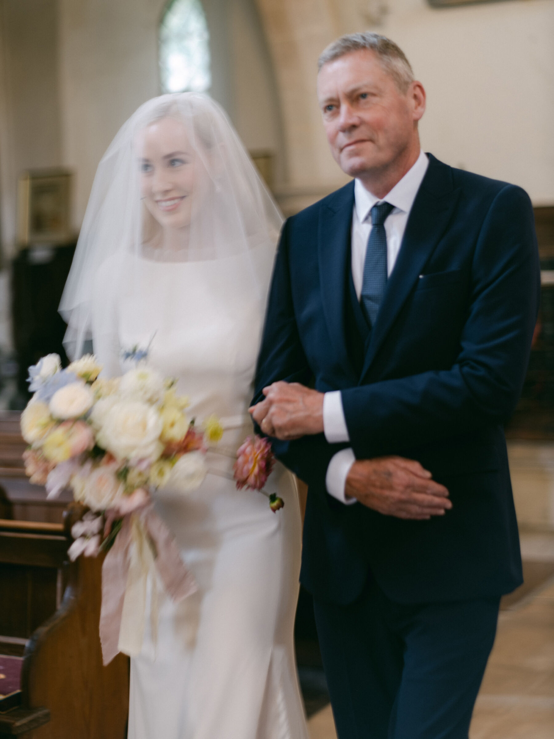 Annie & Harry's Effortlessly Glamorous, Colourful & Modern Double Wedding Weekend 7 Elegant modern bride arm in arm with her father (in a blue suit) with a single tier short veil as she walks down the aisle and smiles at her groom. Katie Julia Photography.