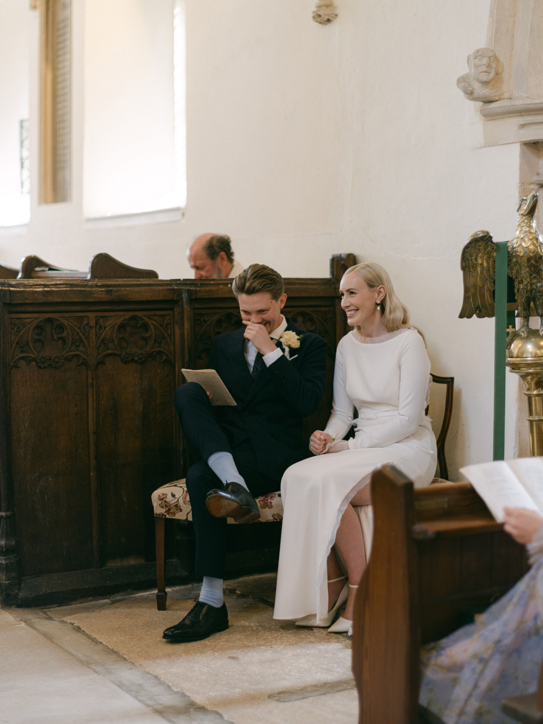 Annie & Harry's Effortlessly Glamorous, Colourful & Modern Double Wedding Weekend 8 Modern bride and groom seated during and giggling during their church ceremony. Groom in pale blue socks. bride in long sleeve thigh slit wedding dress by Stephanie Allin. Katie Julia Photography.