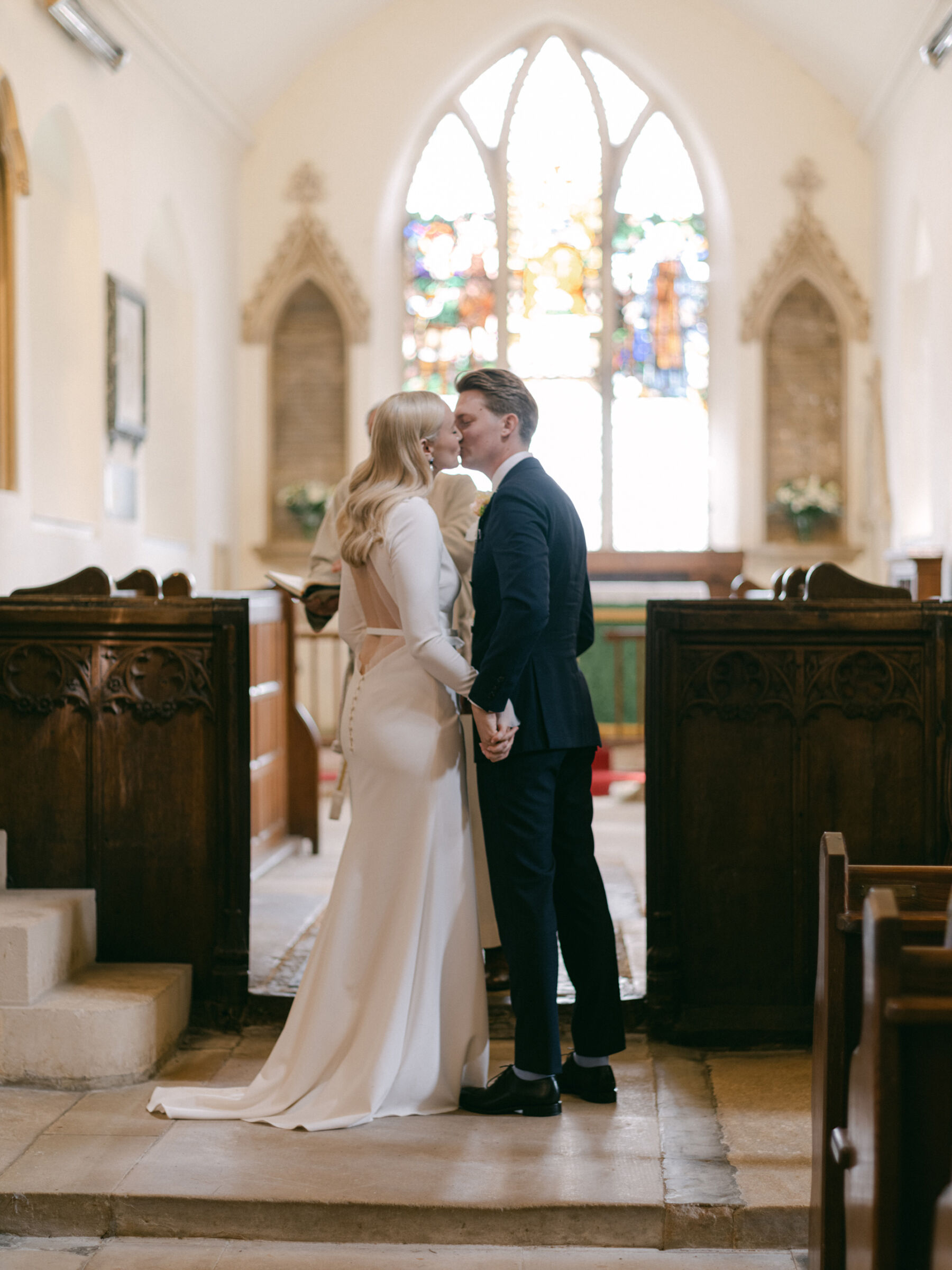 Annie & Harry's Effortlessly Glamorous, Colourful & Modern Double Wedding Weekend 9 Light and airy photography of bride and groom kissing in church. Natural light streaming through the church window in the background. The bride wears a low back bespoke dress by Stephanie Alline. Katie Julia Photography.