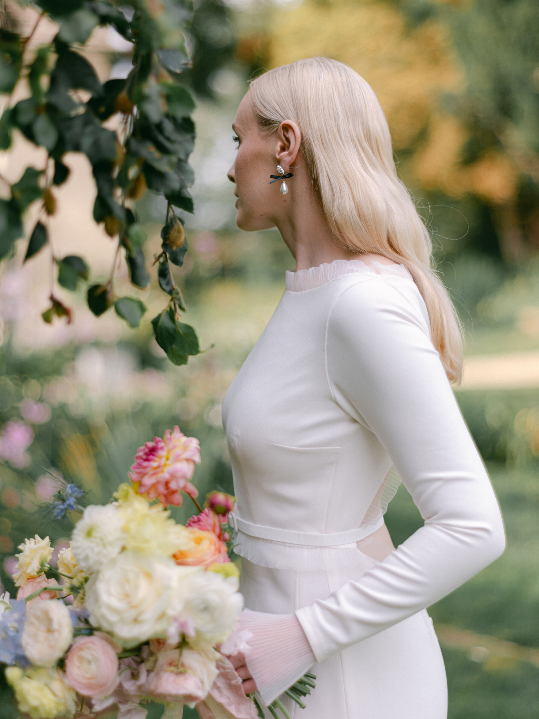 Annie & Harry's Effortlessly Glamorous, Colourful & Modern Double Wedding Weekend 4 Beautiful bride with long blond hair looking away from the camera to reveal pretty pearl earrings. She wars a bespoke Stephanie Allin wedding dress and carries an elegant light pastel colour bouquet in predominantly yellow shades. Katie Julia Photography.
