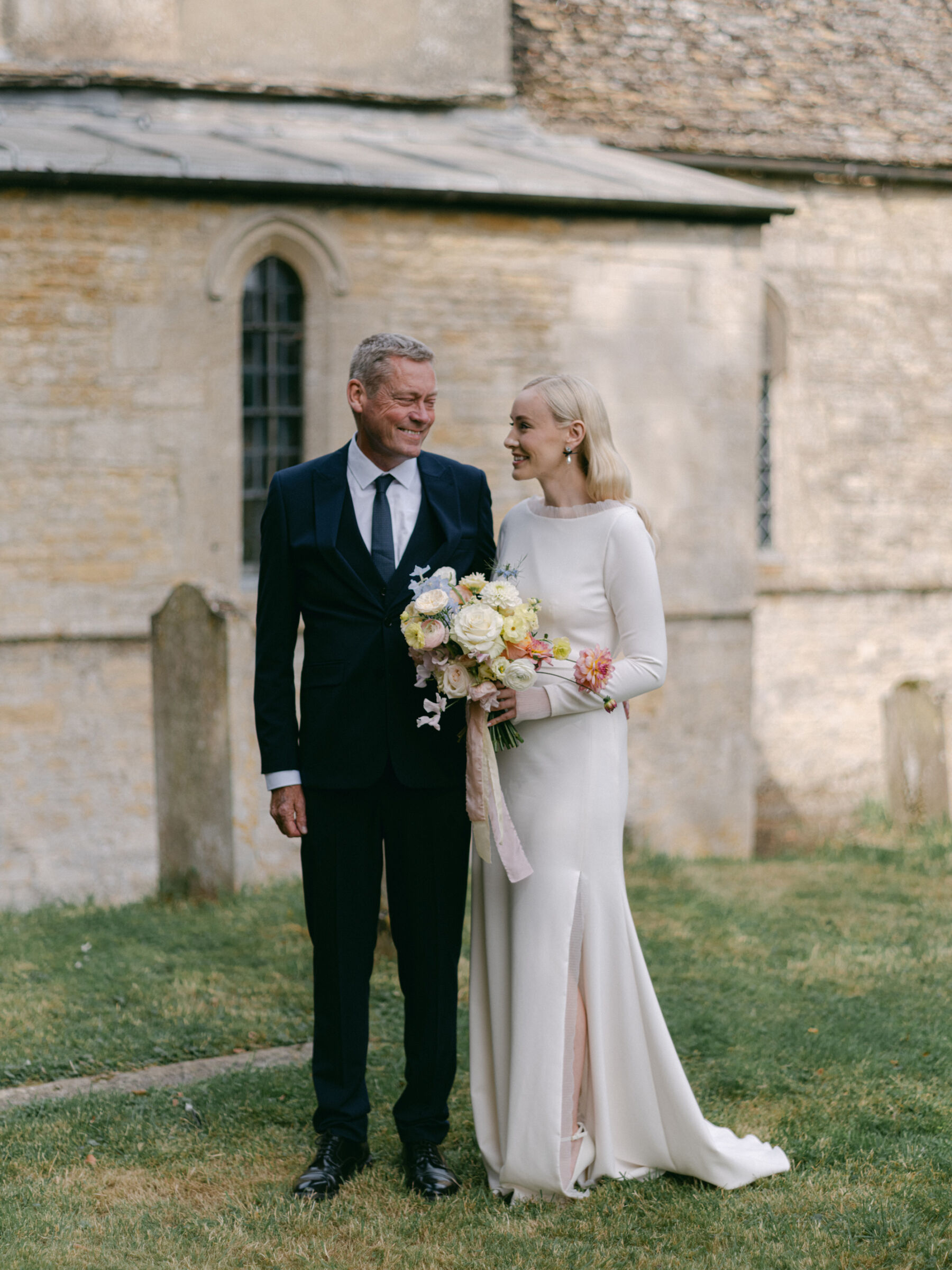 Annie & Harry's Effortlessly Glamorous, Colourful & Modern Double Wedding Weekend 3 Bride and father of the bride standing outside the church moments before the ceremony. They are smiling at each other - the elegant modern bride carries a big bouquet of mostly yellow summer flowers with a very pale pink silk ribbon. She wears a long sleeved dress with frilly tulle cuffs and a thigh slit by Stephanie Allin. Katie Julia Photography.
