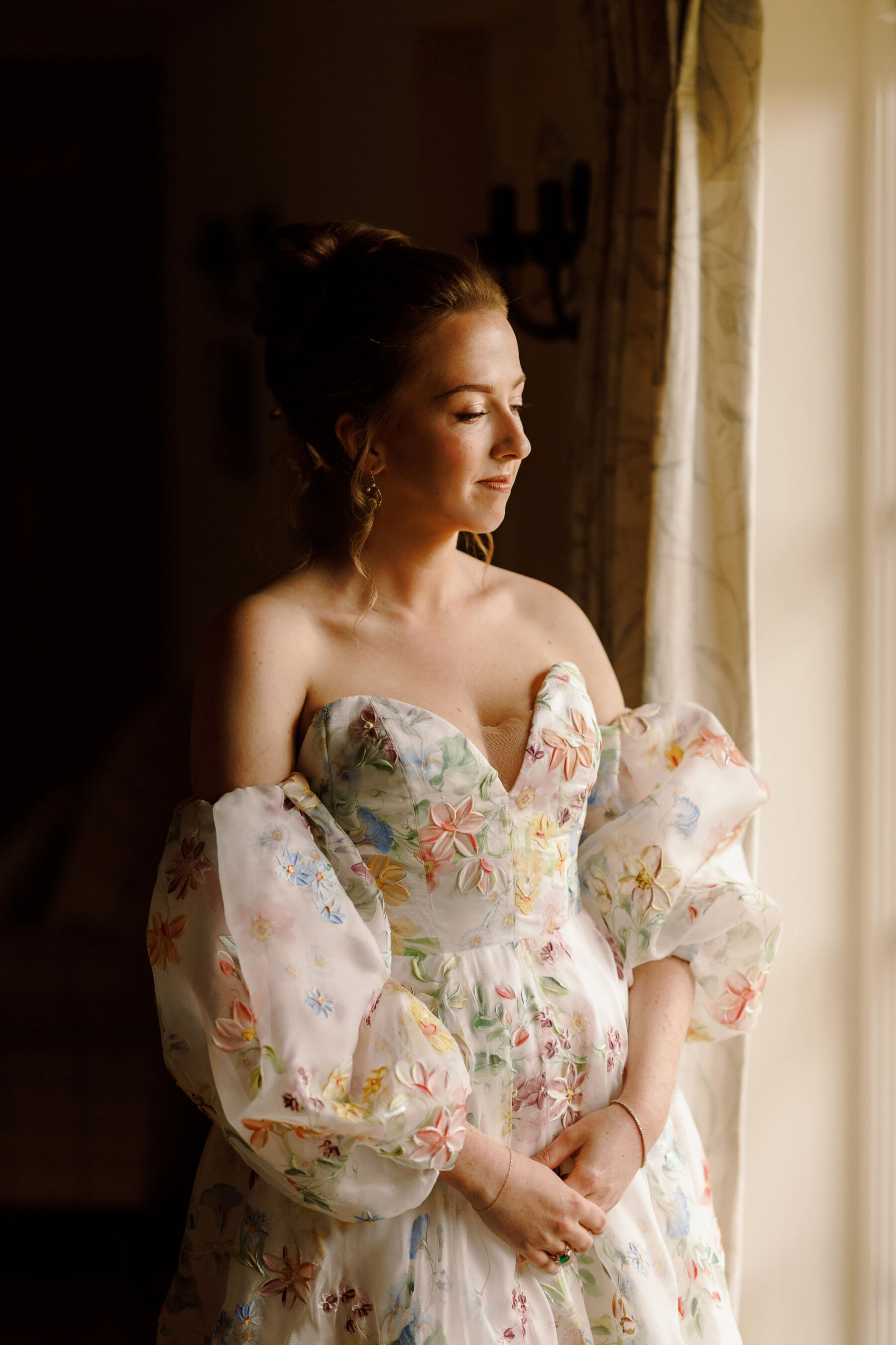 Bride wearing a floral wedding dress by House of Savin with detachable sleeves. She is standing in natural window light and wears a romantic up do in her hair.