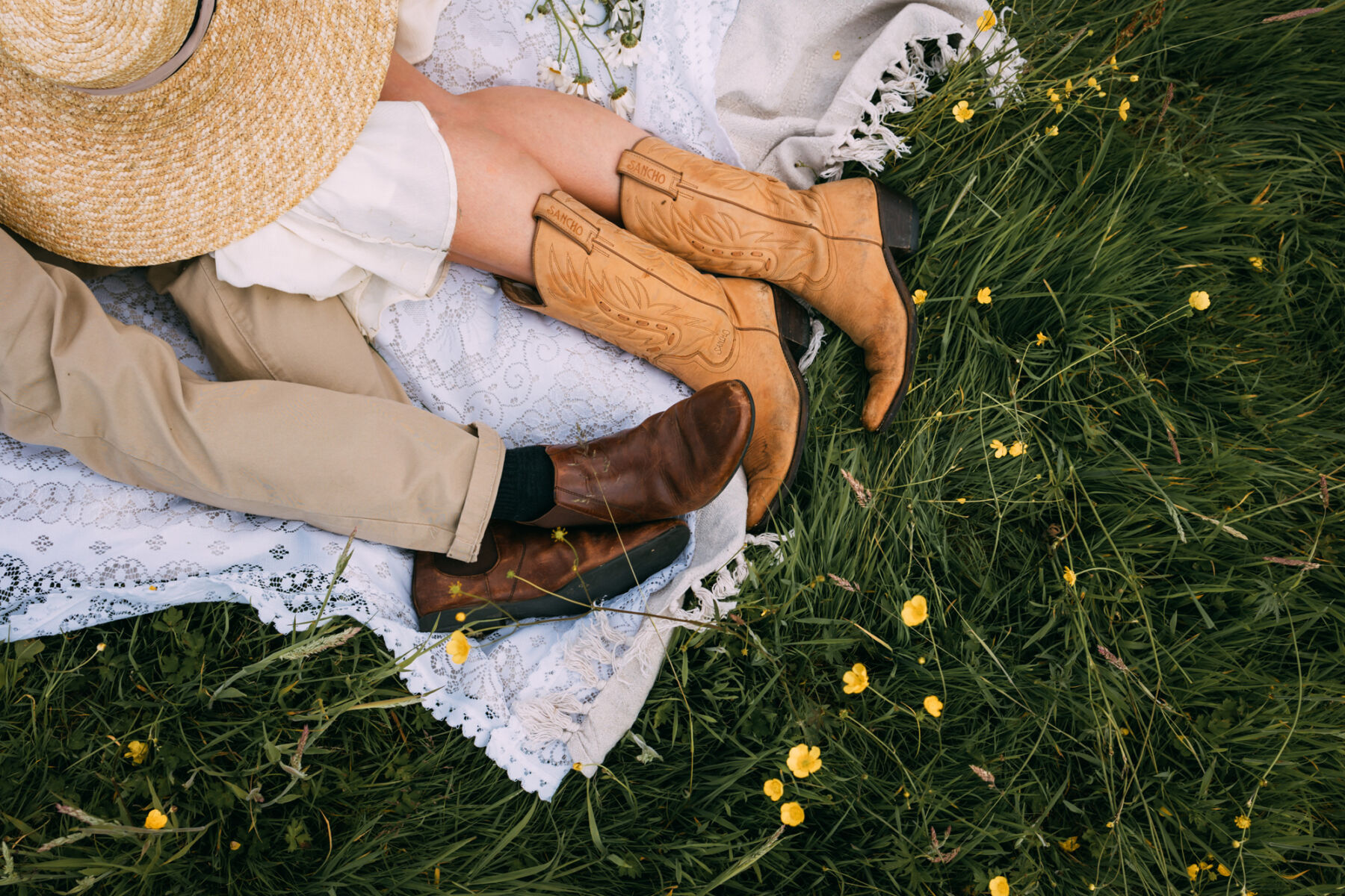 Wickerwood Farm, an Eco Friendly, Glamping Wedding Venue in East Sussex 17 Birds eye view of the legs of a bride in cowboy boots as she cuddles with her groom. A strawhat is nearby and they are lying in a field full of buttercups.