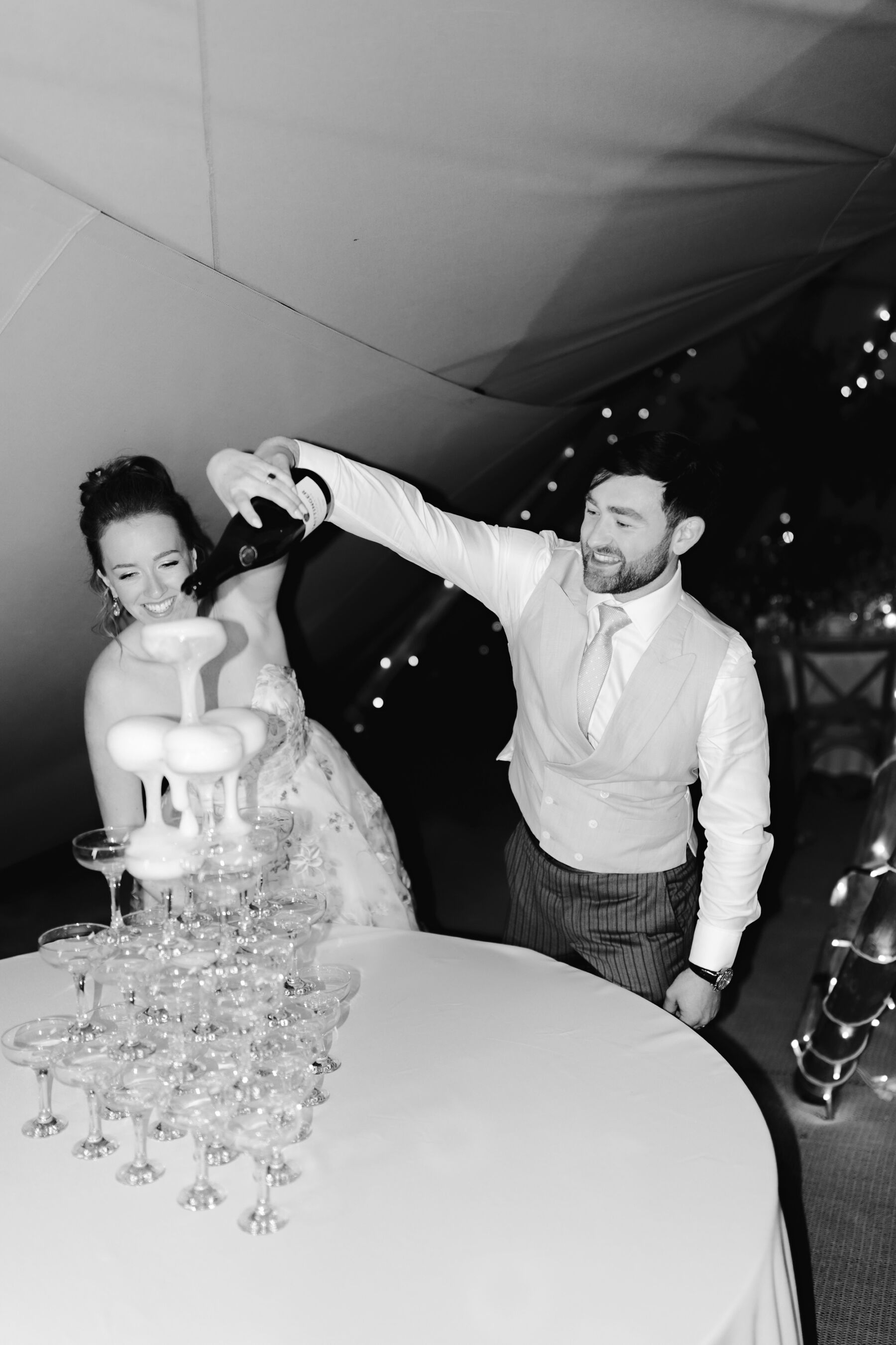 Bride and groom pouring into the top glass of a champagne tower - the champagne bubbles are throthing over the top of the glasses.