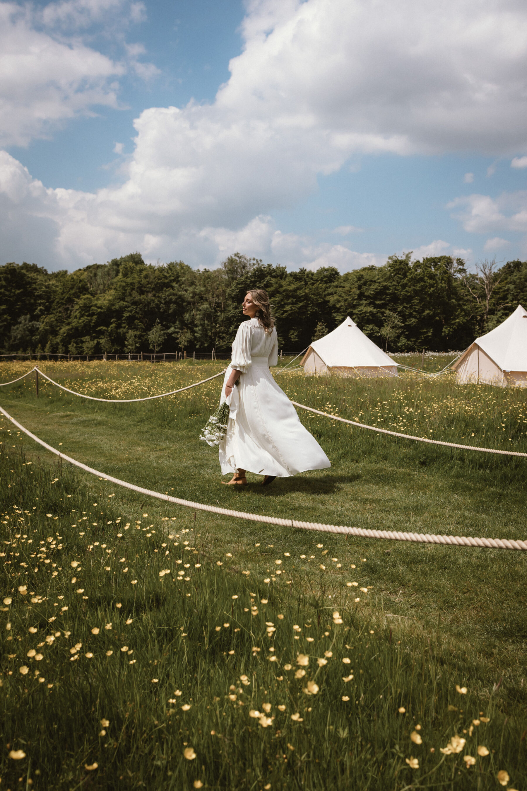 Wickerwood Farm, an Eco Friendly, Glamping Wedding Venue in East Sussex 4 Bride wandering through a field full of buttercups with bell tents. She carries a bouquet of large daisies and wears a Free People wedding dress.