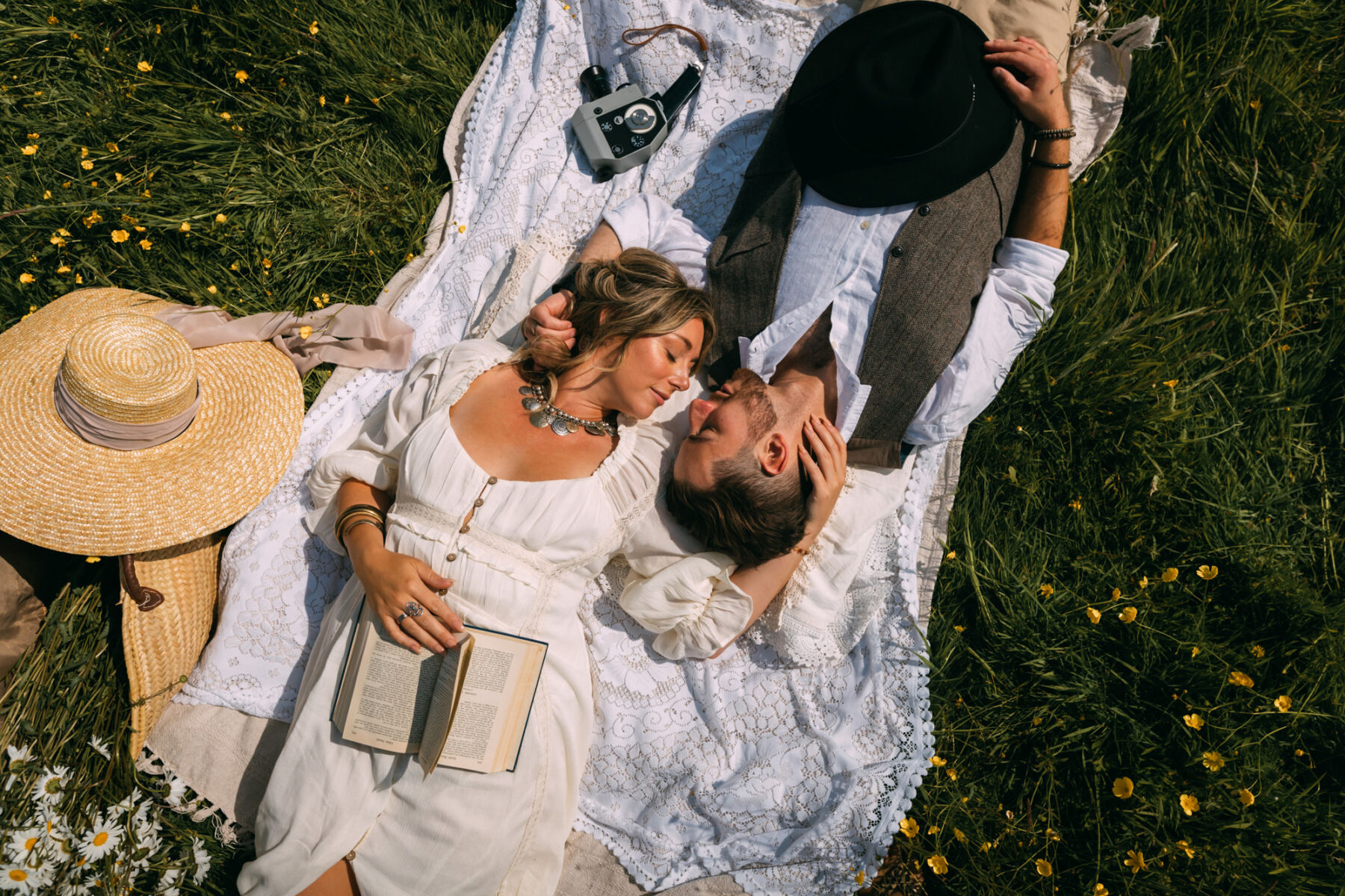 Wickerwood Farm, an Eco Friendly, Glamping Wedding Venue in East Sussex 21 Birdseye view of a romantic scene of a bride and groom lying on a blanket in a field full of buttercups, they are lying in opposite directions but facing one another's face and tenderly cradling each other's heads. A straw hat is nearby. Wickerwood Farm wedding venue, East Sussex.