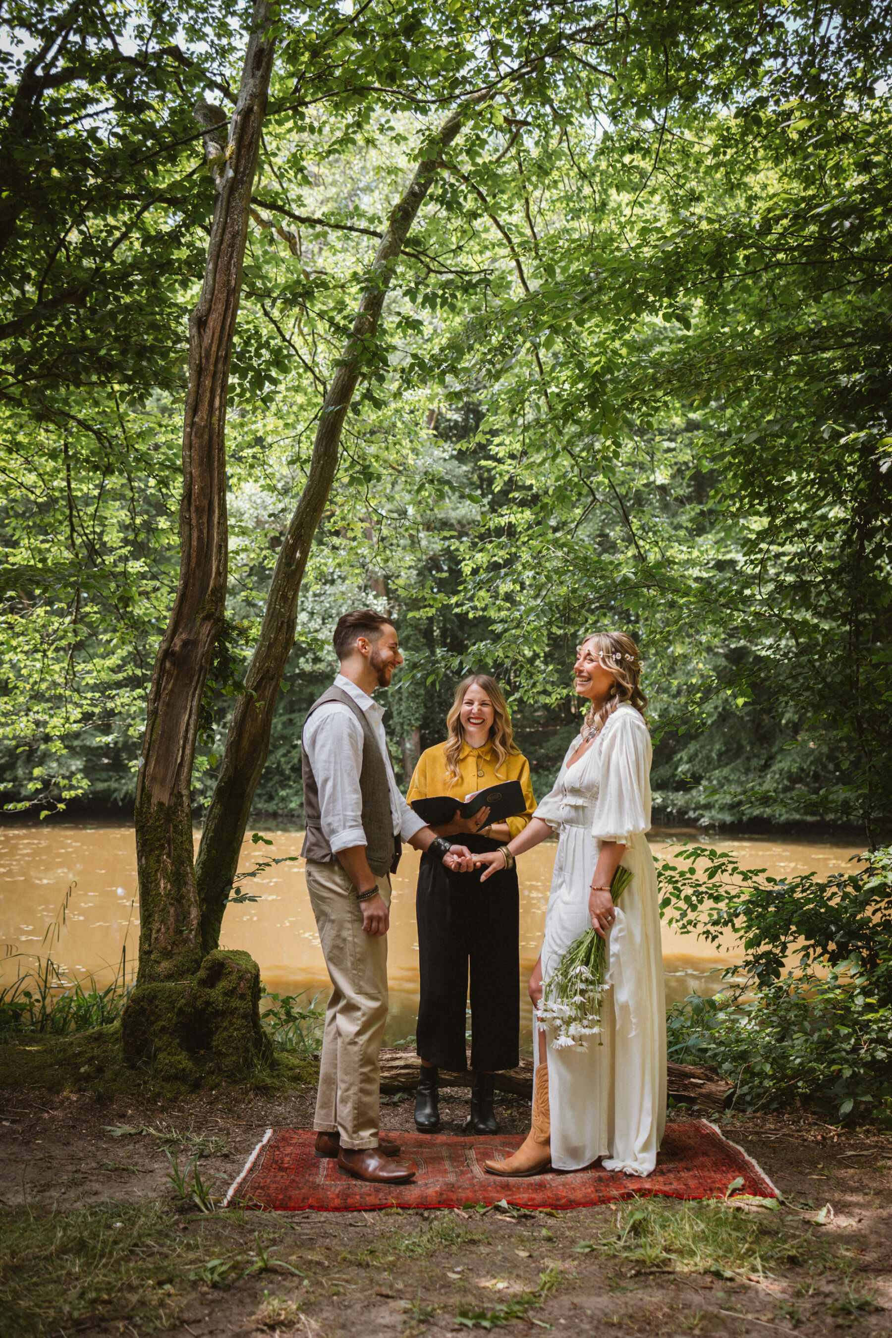 Wickerwood Farm, an Eco Friendly, Glamping Wedding Venue in East Sussex 5 Bride and groom standing on a Persian matt by the river, as they exchange wedding vows with a happy smiling Celebrant standing nearby.