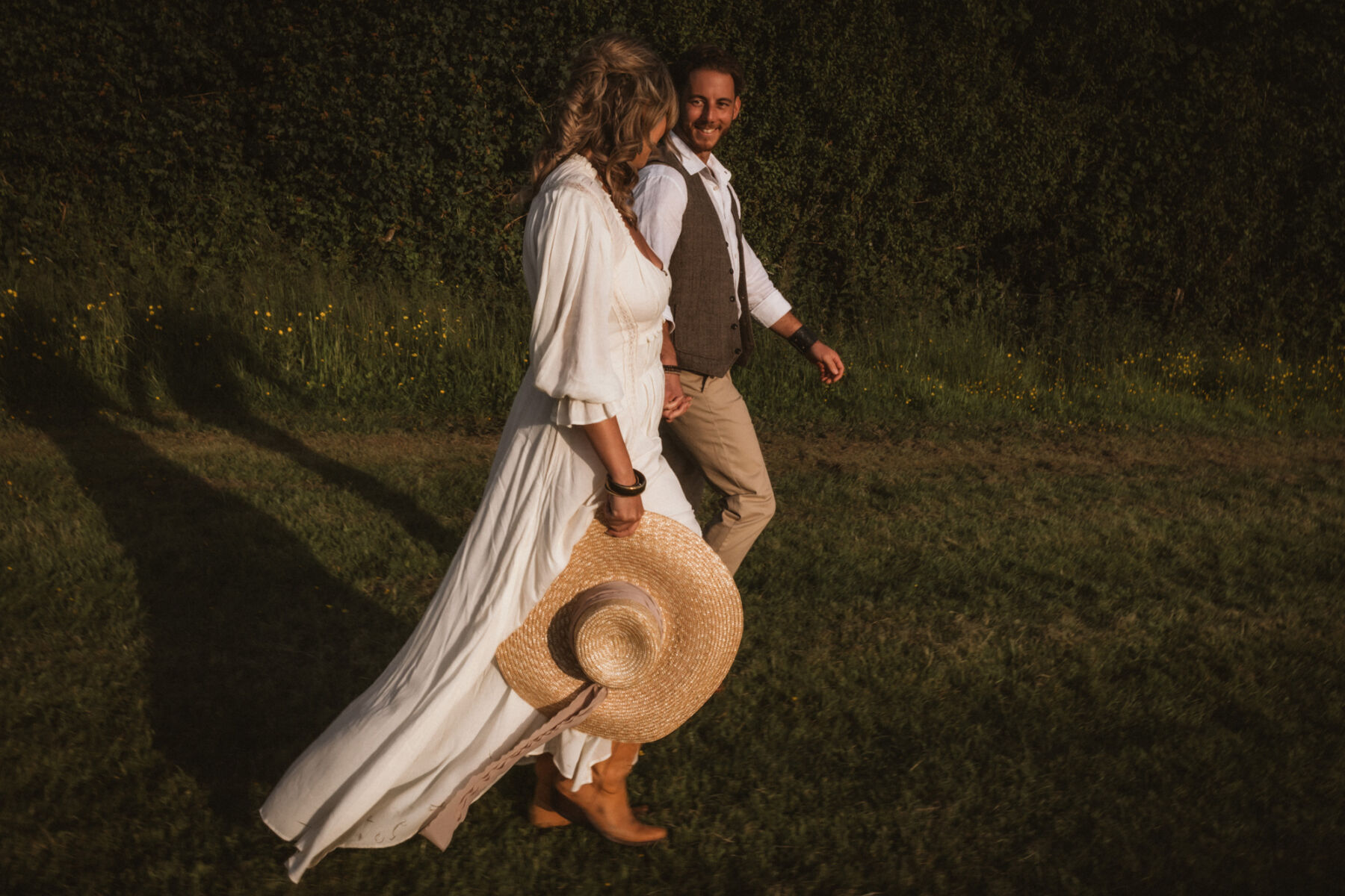 Wickerwood Farm, an Eco Friendly, Glamping Wedding Venue in East Sussex 27 Bride and groom holding hands and wandering through a field at golden hour. The bride wears a Free People wedding dress and carries a straw hat in her hand.