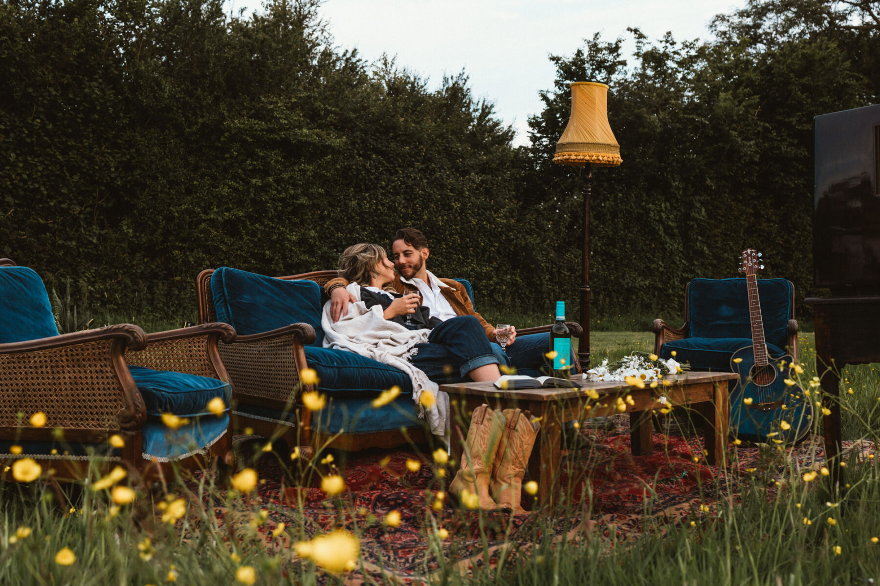 Wickerwood Farm, an Eco Friendly, Glamping Wedding Venue in East Sussex 26 Anitque wedding props and furniture in a field, including chairs with blue velvet cushions, tassled lamp and wooden table. Bride and groom are cuddling on a vintage sofa.