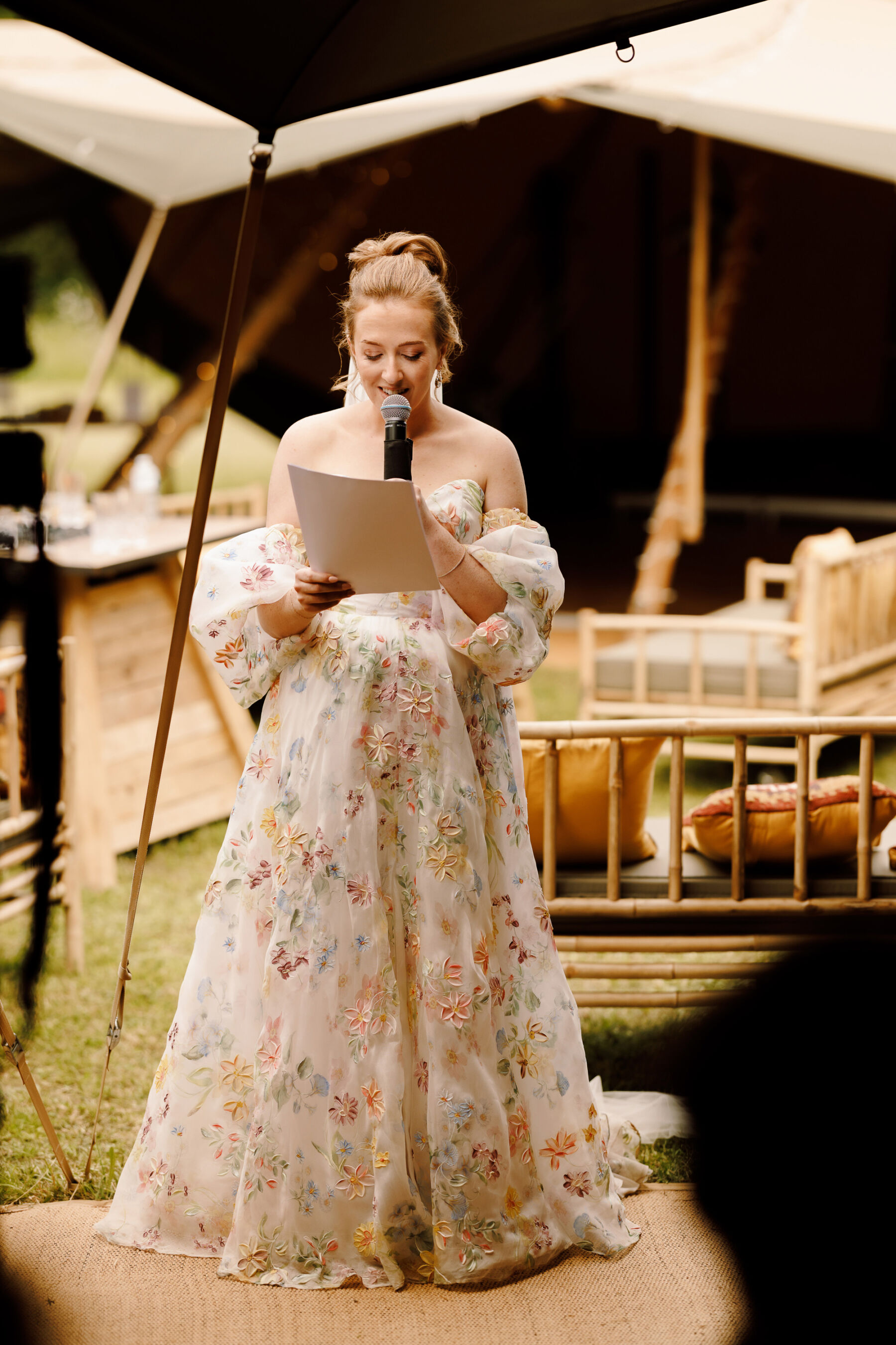 Bride holding a microphone to give a speech on her wedding day. She wears a floral wedding dress by House of Savin.