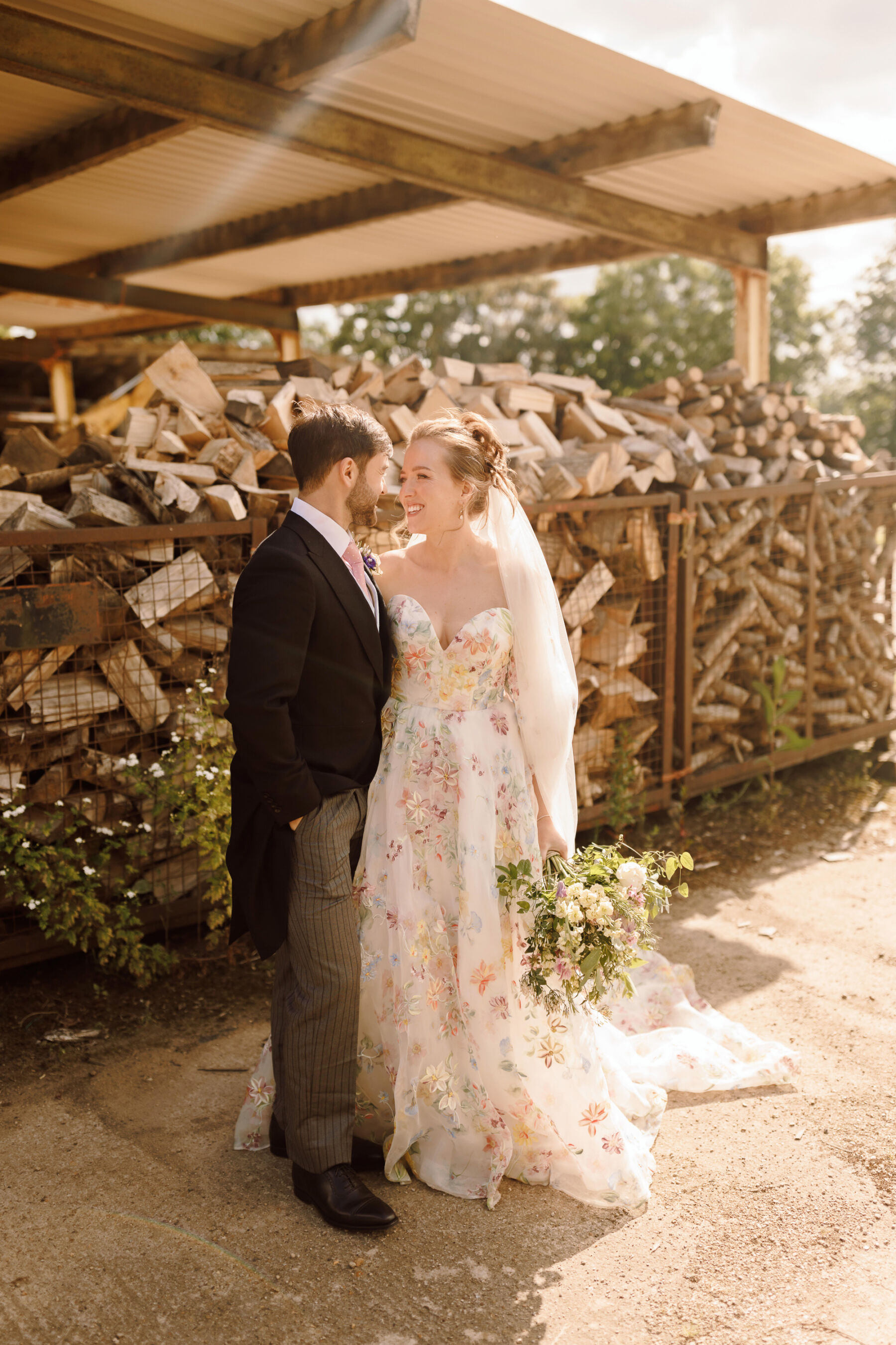 Bride and groom portrait in the sunshine. The bride wears a floral wedding dress by House of Savin with a long veil and carries an elegant summer bridal bouquet. Groom wears a traditional morning suit with pale blue waistcoast and pink tie.