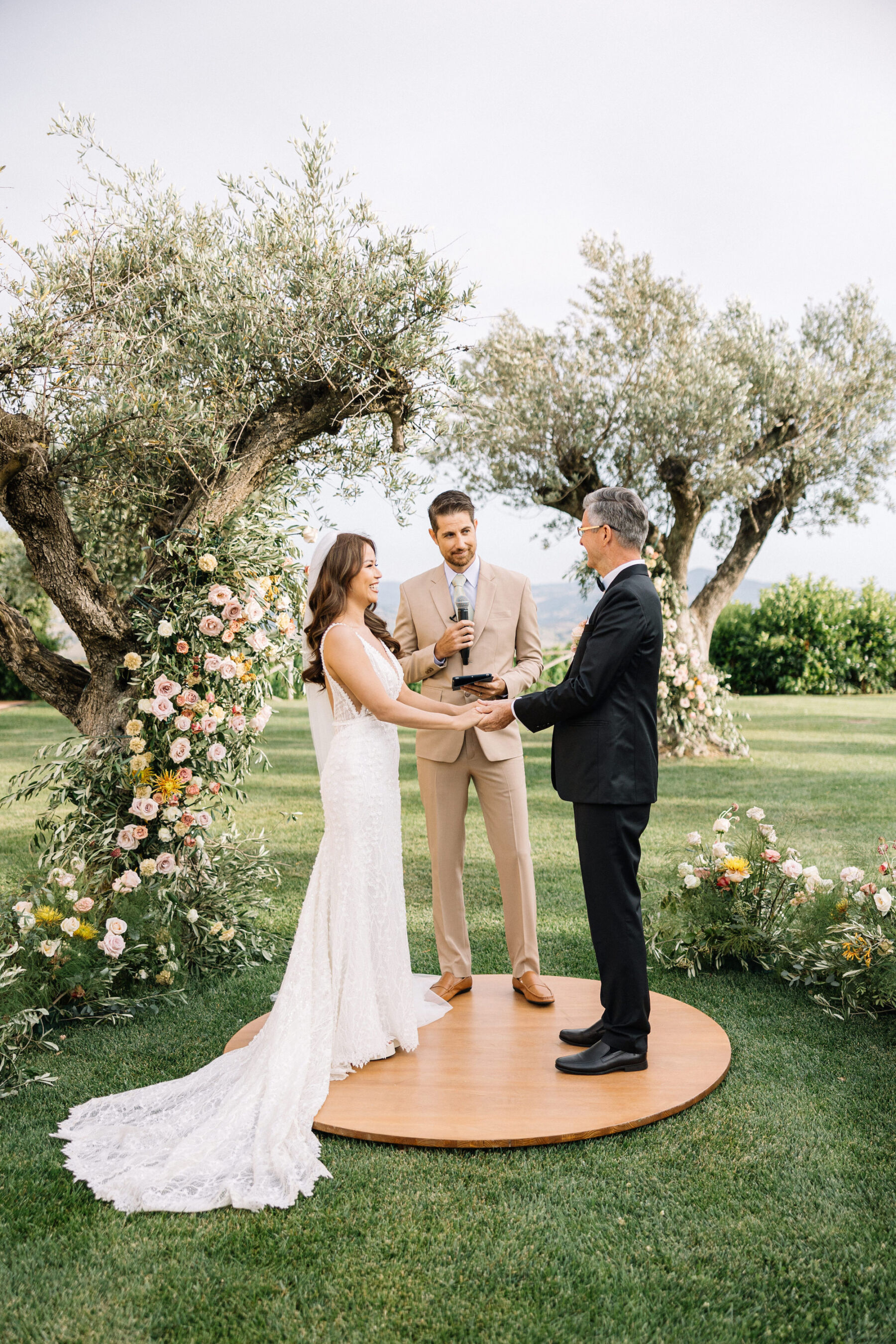 A Professional Wedding Planner's Wedding at Colle Massari in Tuscany, Italy 22 Bride and groom standing on a small round wooden platform with their wedding celebrant as they exchange vows during their wedding ceremony. They are surrounded by olive trees and pastel flowers.