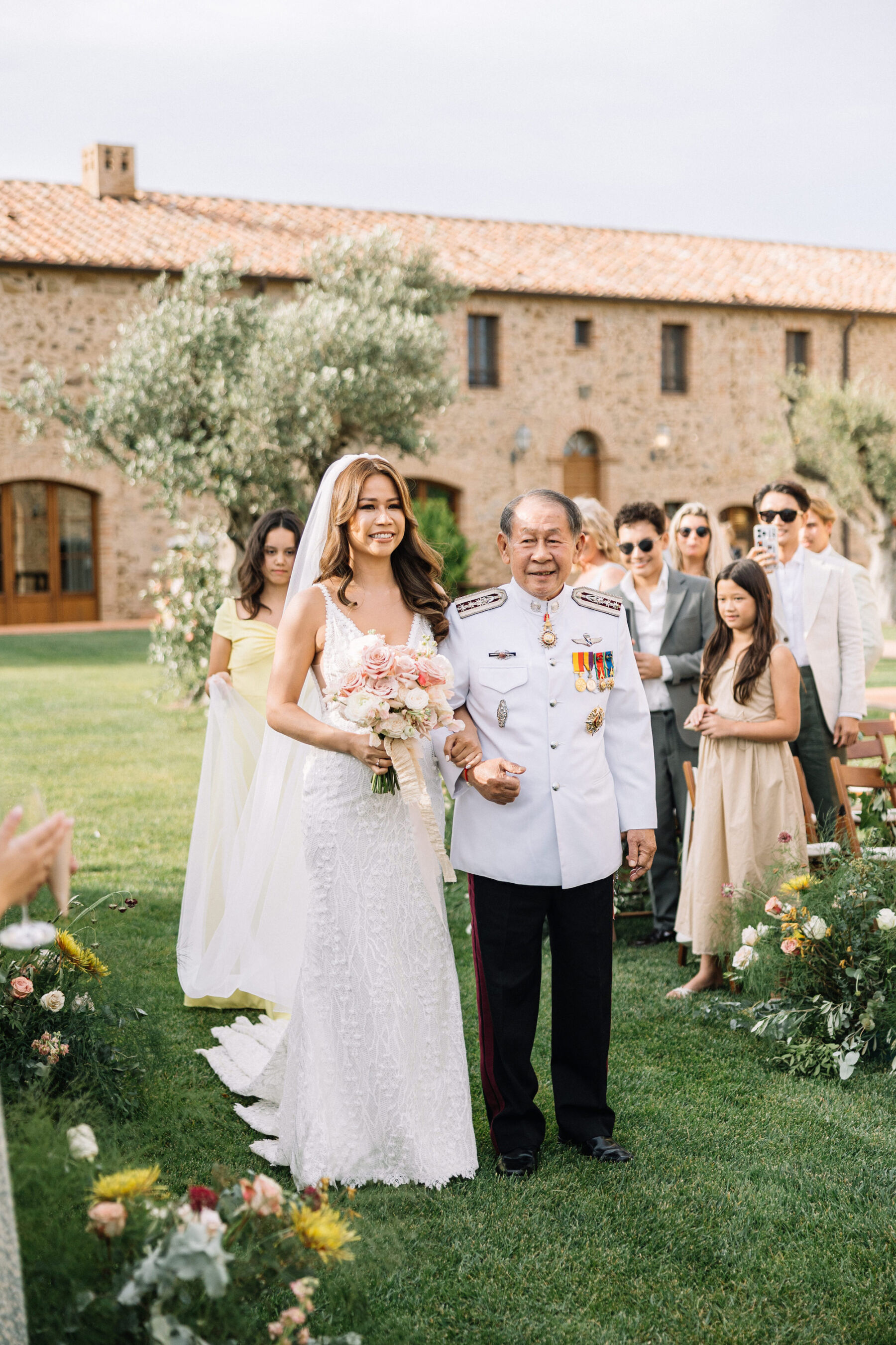 A Professional Wedding Planner's Wedding at Colle Massari in Tuscany, Italy 20 Thai bride and her father walking down the aisle for her wedding day. Olive trees in the backrgound. She carries a pastel wedding bouquet.