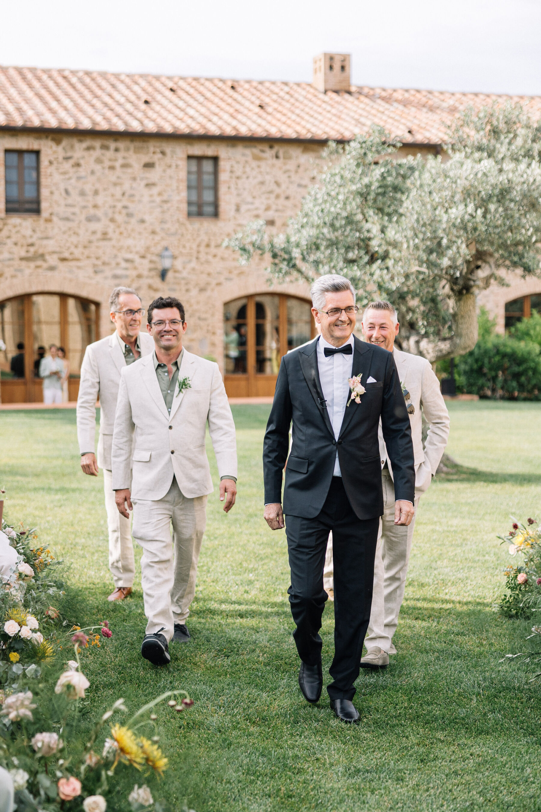 A Professional Wedding Planner's Wedding at Colle Massari in Tuscany, Italy 19 Groom in elegant black tie walking down the aisle of his outdoor wedding in Tuscany. He is accompanied by groomsmen in relaxed beige linen suits.