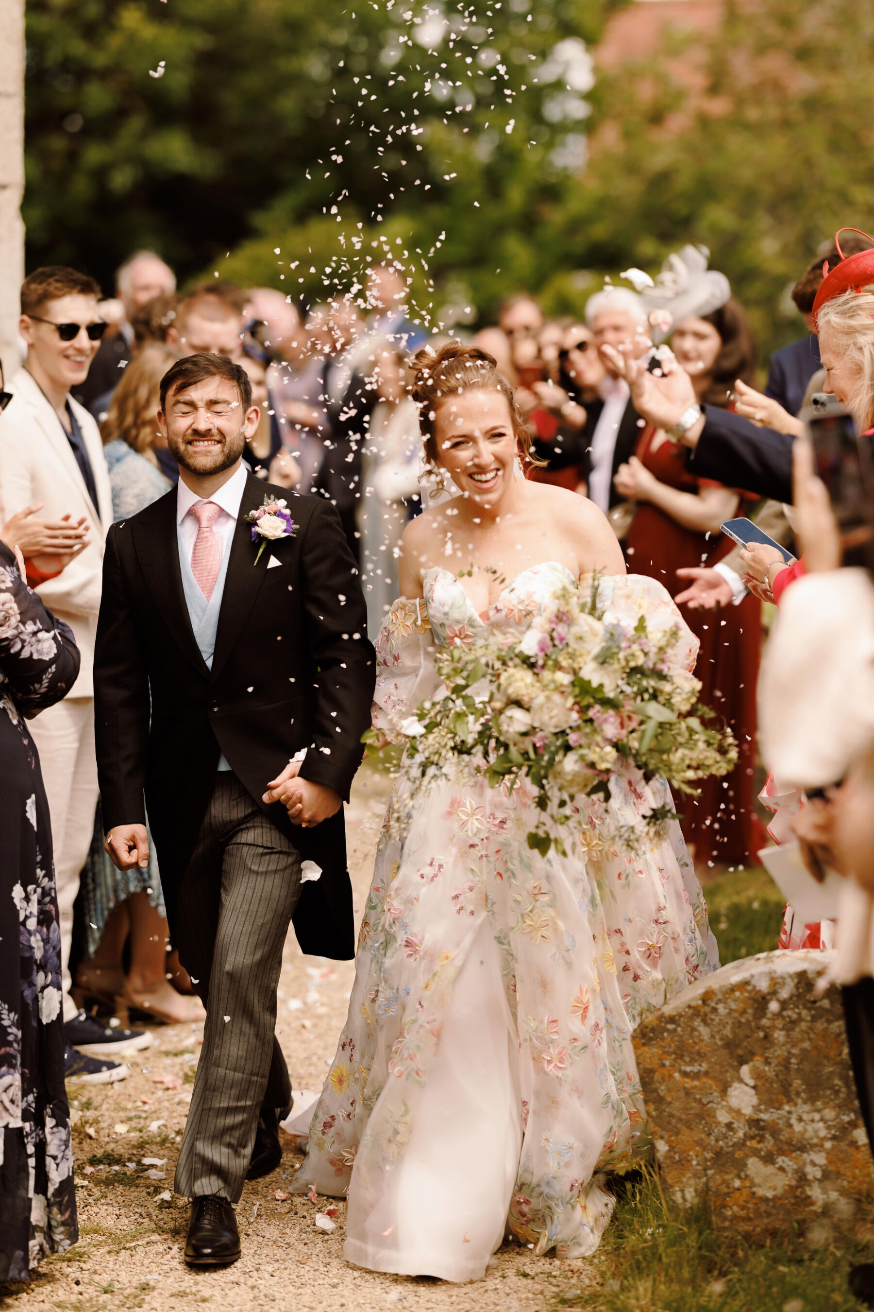 Bride and groom walking through confetti. The bride wears a floral wedding dress by House of Savin and the groom wears a morning suit with a pale blue waistcoat and pink tie.