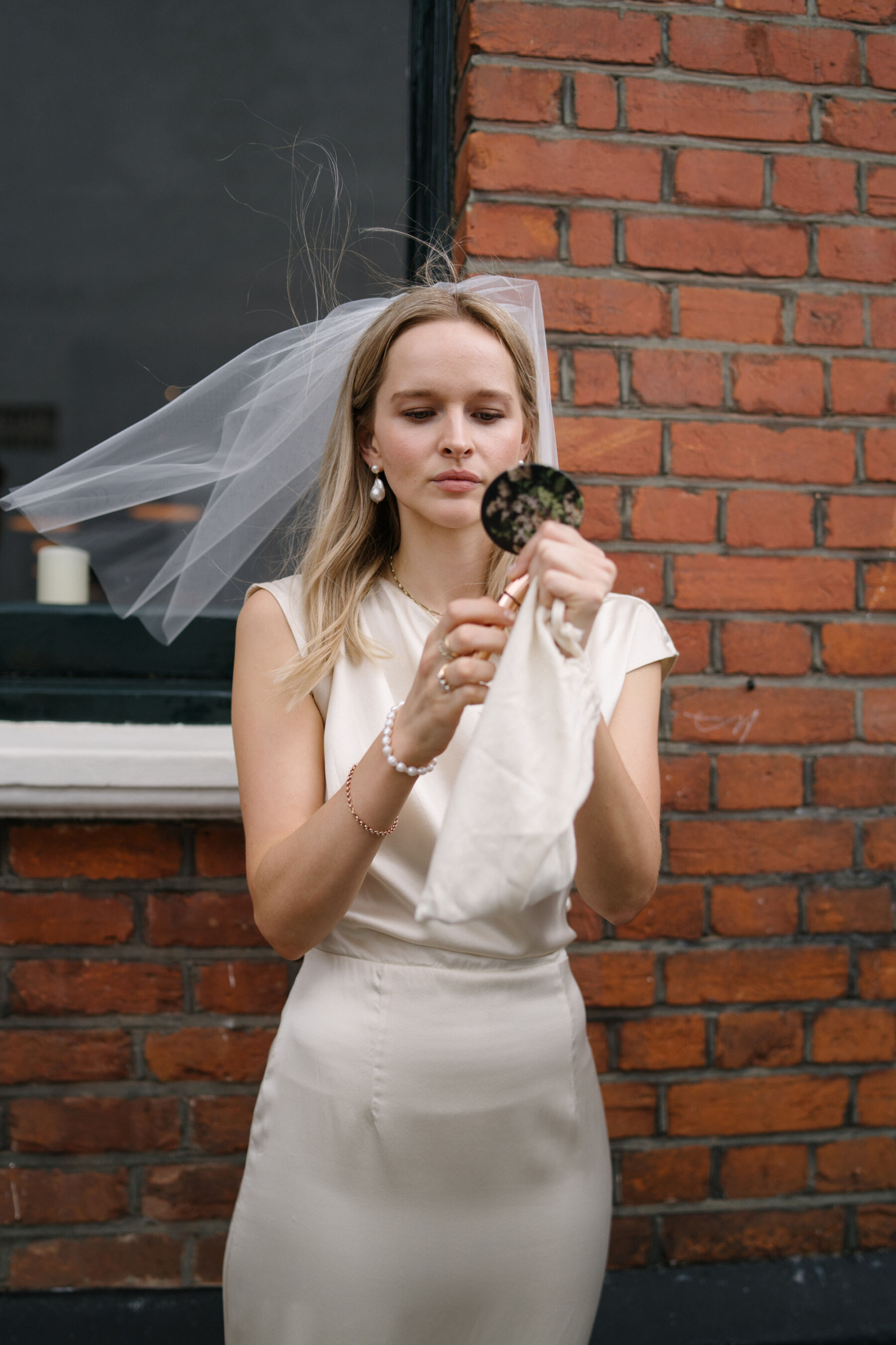 Relaxed, Romantic & Organic: Elena and Oliver's Cambridge Cottage Wedding at Kew Gardens 16 Modern bride checking her makeup in the mirror, her short veil caught in the breeze. She wears a Kamperett wedding dress from The Fall Bride and wears pearl earrings.