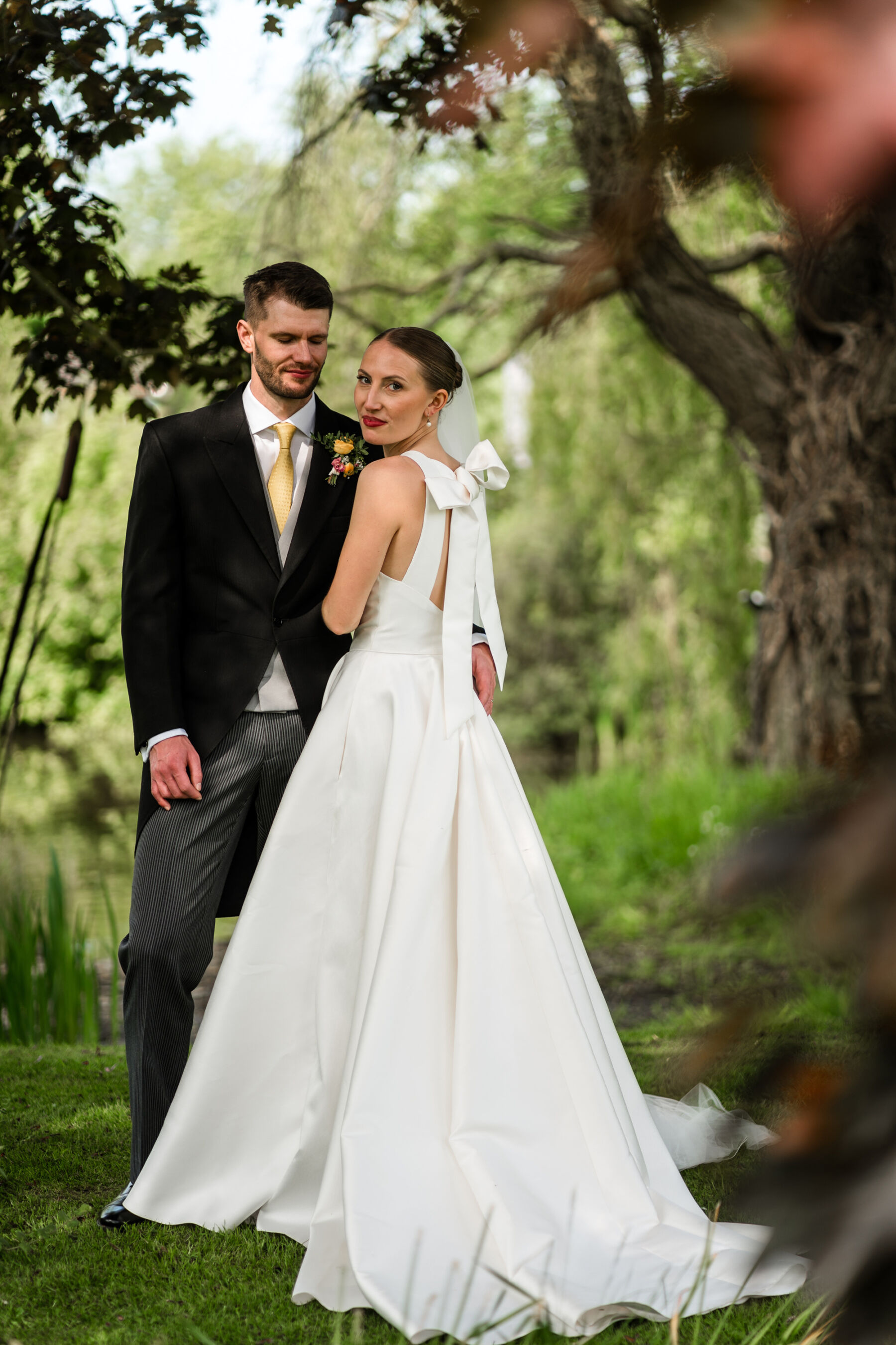 Alice Wore Oak by Halfpenny London For Her Charming Spring Marquee Wedding in Suffolk 25 Bride turning back to look at the camera, showing the back of her dress, the Oak by Halfpenny London, which features a large bow tied at the top of her back. Her Groom wears traditional morning suit and yellow tie.