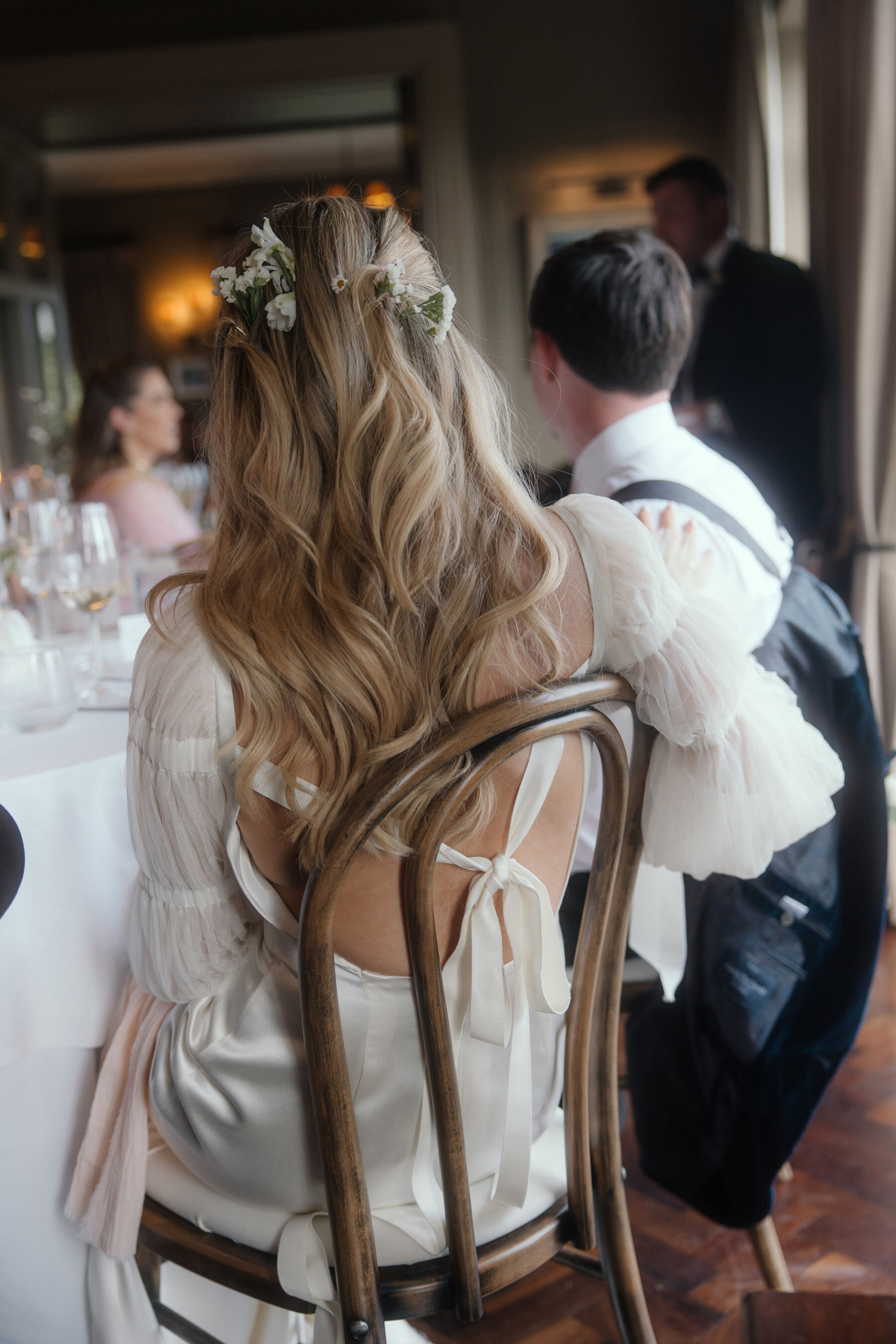 Romantic Ballynahinch Castle Wedding: Ciara in Danielle Frankel 30 Bride with Feverfew in her hair and wearing a Danielle Frankel bridal gown as she sits on a wooden chair at her wedding reception inside Ballynahinch Castle, Ireland.