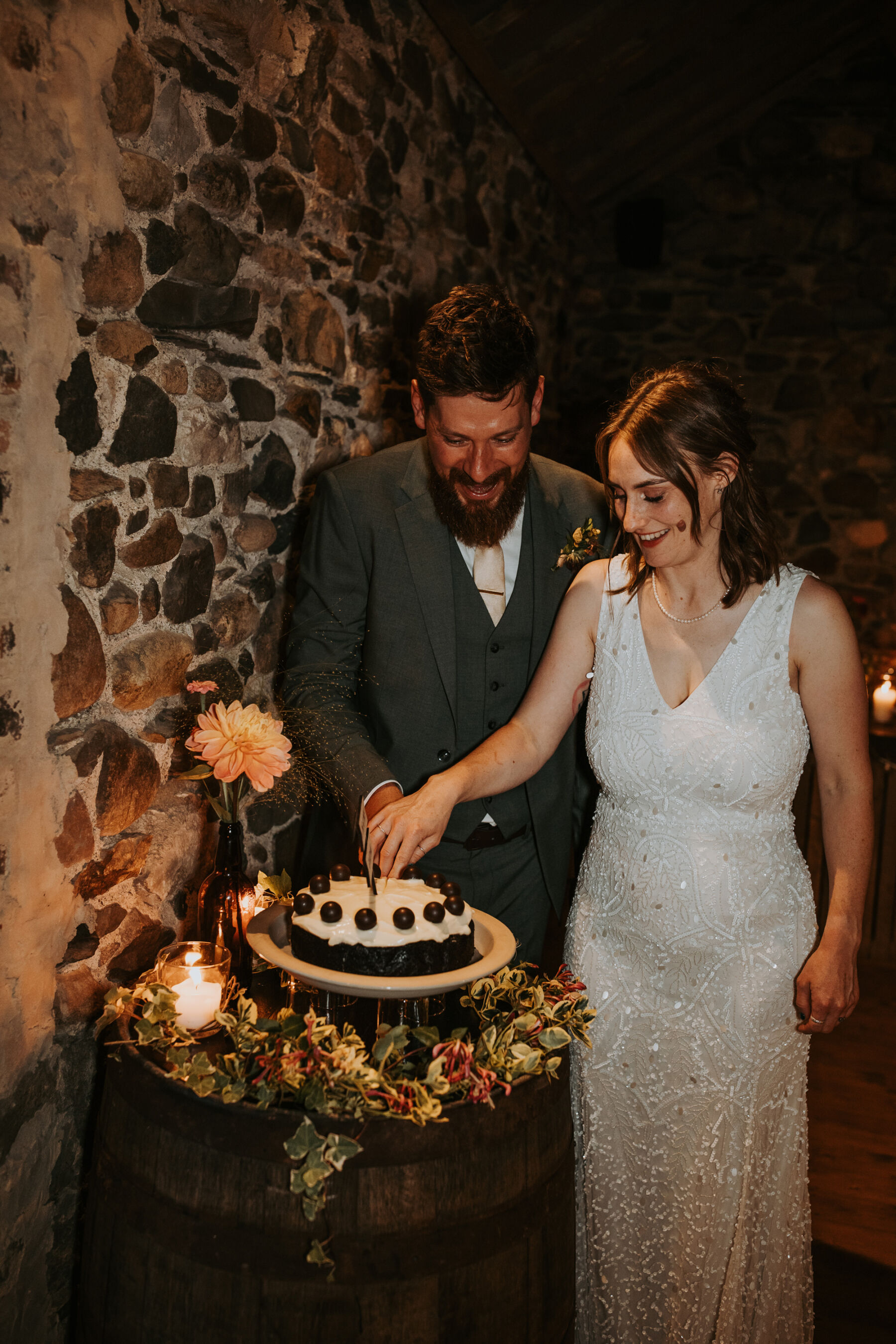 Sophie & Hal's Joyful, Colourful & Sustainable DIY Barn Wedding at Low Hall The Lakes 50 Bride and groom holding the cake knife together to make the first cut. Single tier wedding cake in inside a barn with a Dahlia flower neaby.